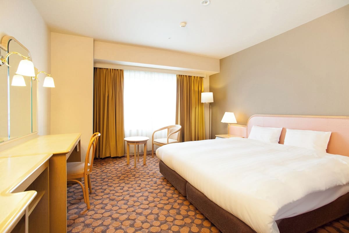 A hotel room with a bed, a desk, a chair, a small table, a lamp, and a window with curtains.