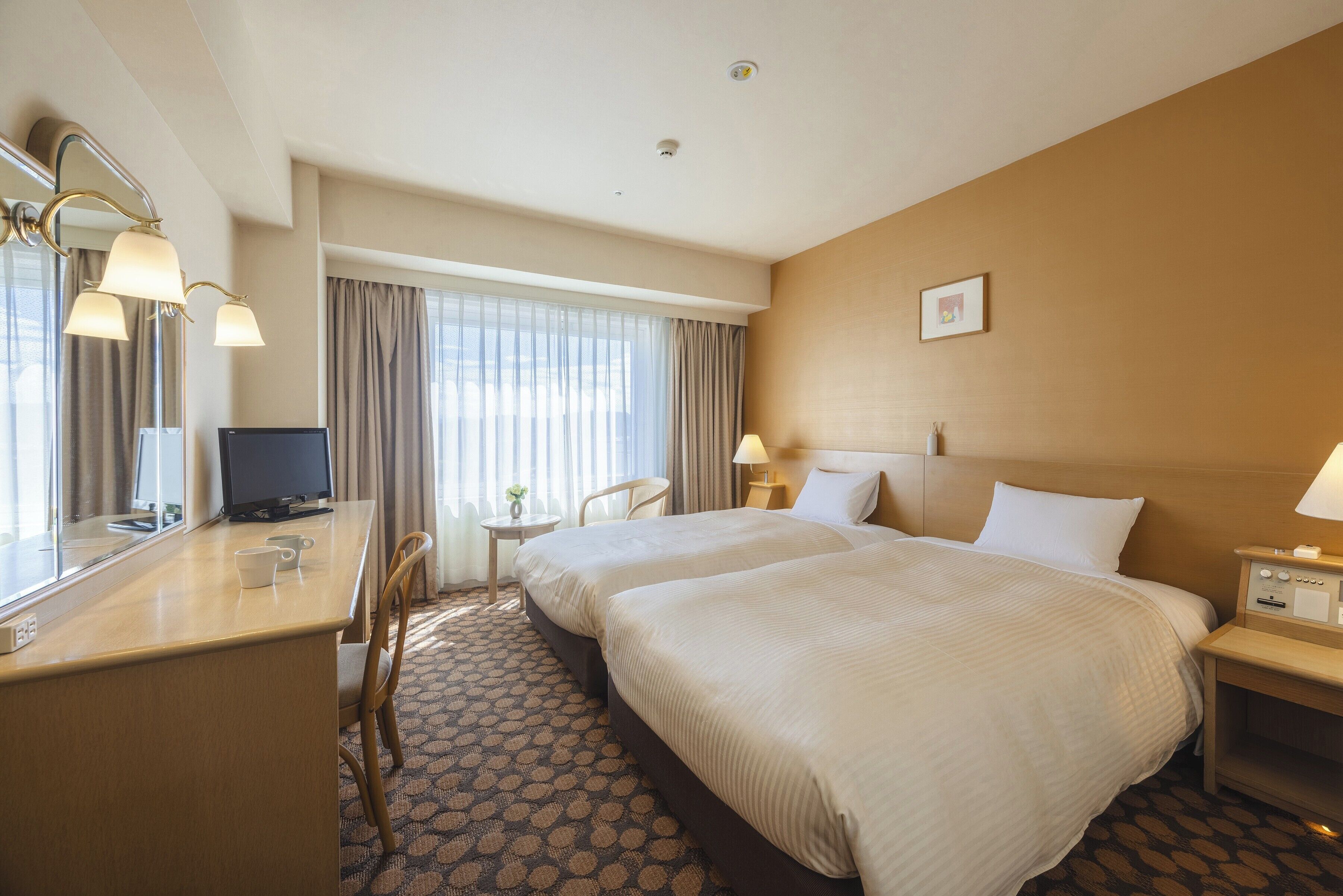 Habitación de hotel con dos camas, un escritorio con computadora, una silla, una mesita de noche con lámpara y ventana con cortinas.