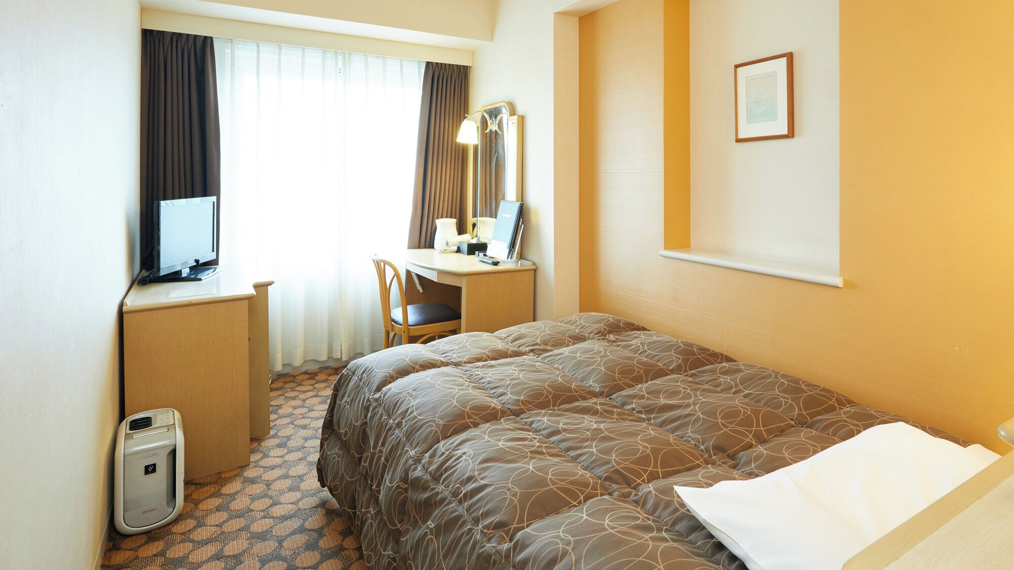 A hotel room with a bed, a desk with a computer, a chair, a desk lamp, and a small trash bin.