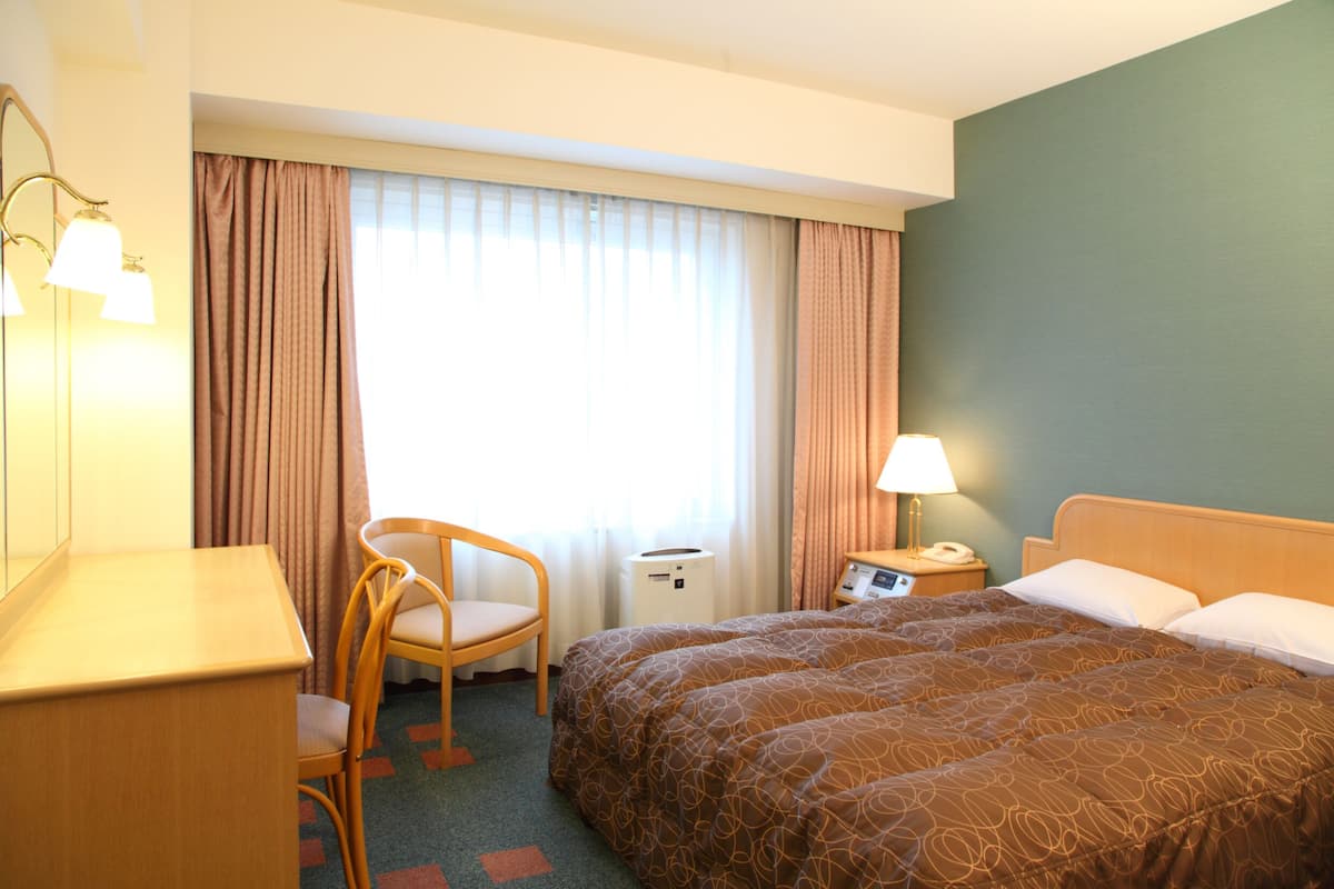 A hotel room with a large bed, a desk, a chair, a nightstand with a lamp, and a window with curtains.