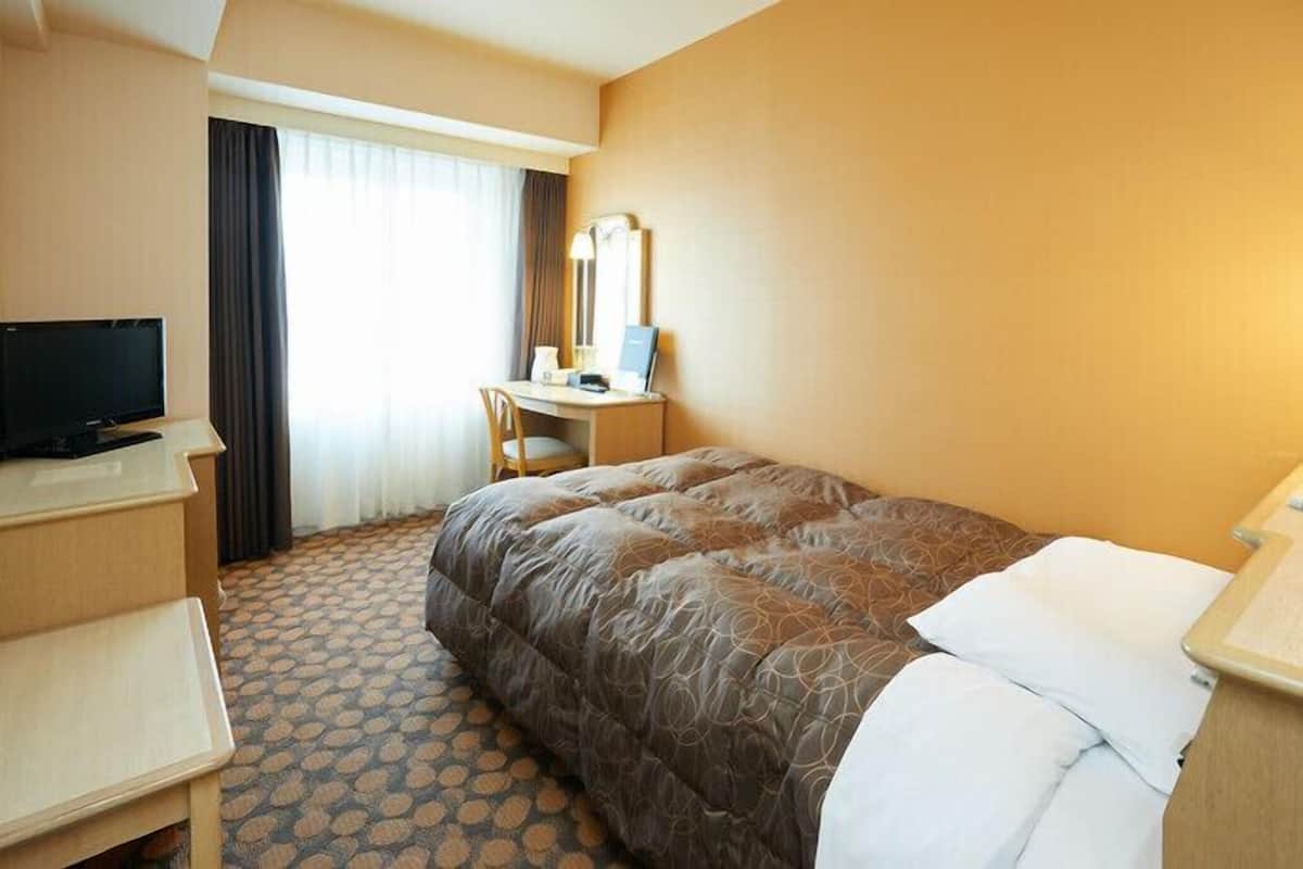 A hotel room with a bed, a desk with a chair, a television, and a window with curtains.