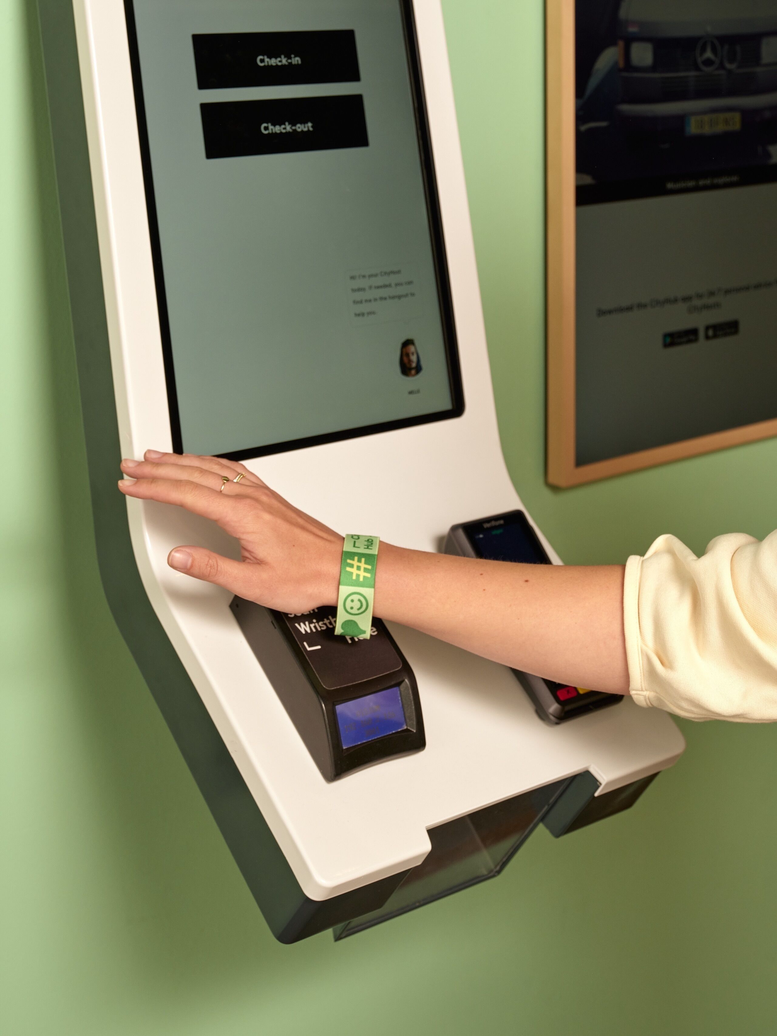 A person using a self-service kiosk with a green wristband, a check-in screen, and a payment terminal.
