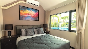 A hotel room with a large bed, two bedside tables with lamps, a painting above the bed, and a window with a view of a pool and palm trees. - Pattaya Hill, Near the City and Beach (Pattaya)