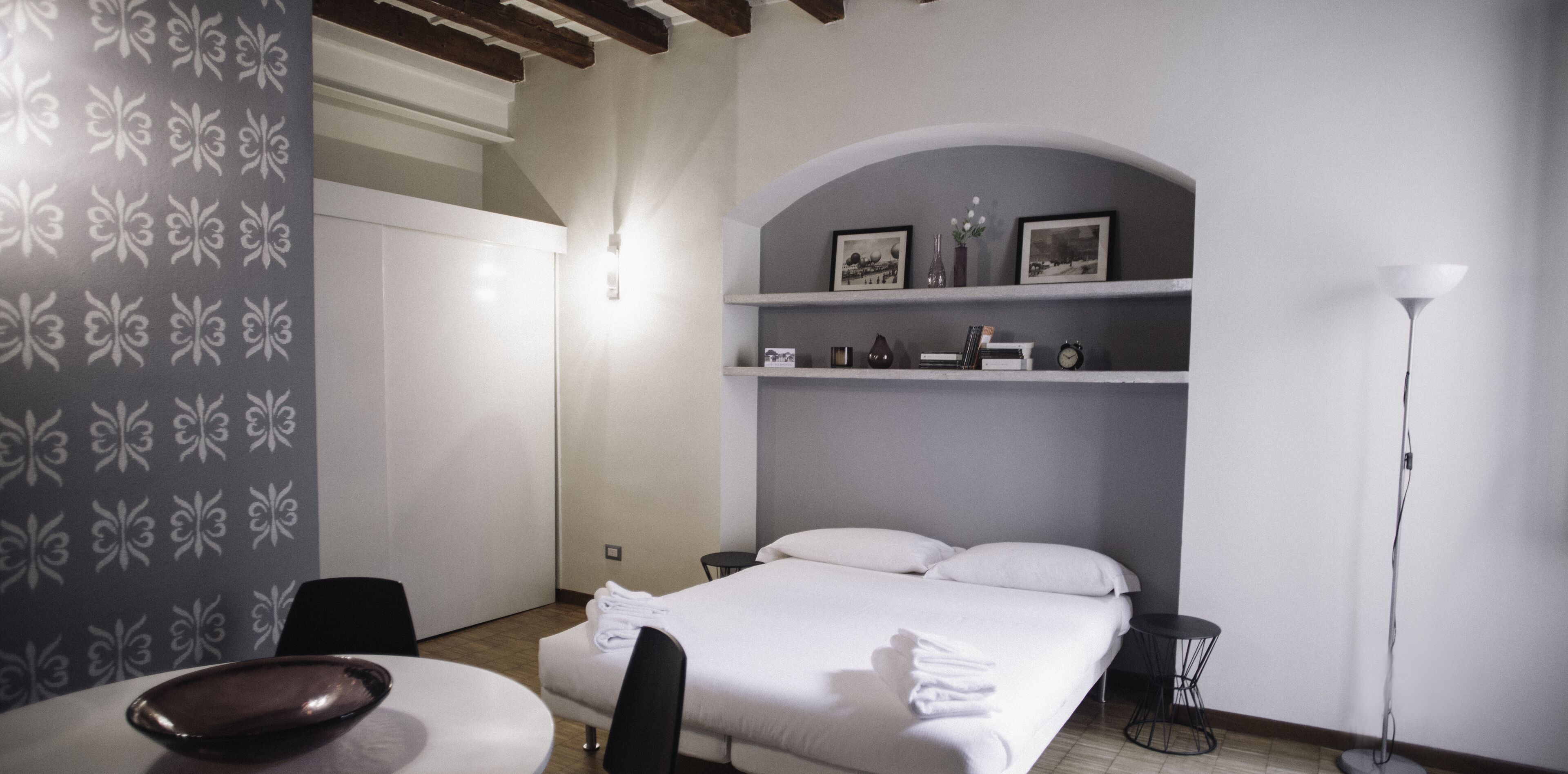 Una camera d'albergo con un letto, una scrivania, una sedia, un tavolino, una lampada e uno scaffale con oggetti decorativi.