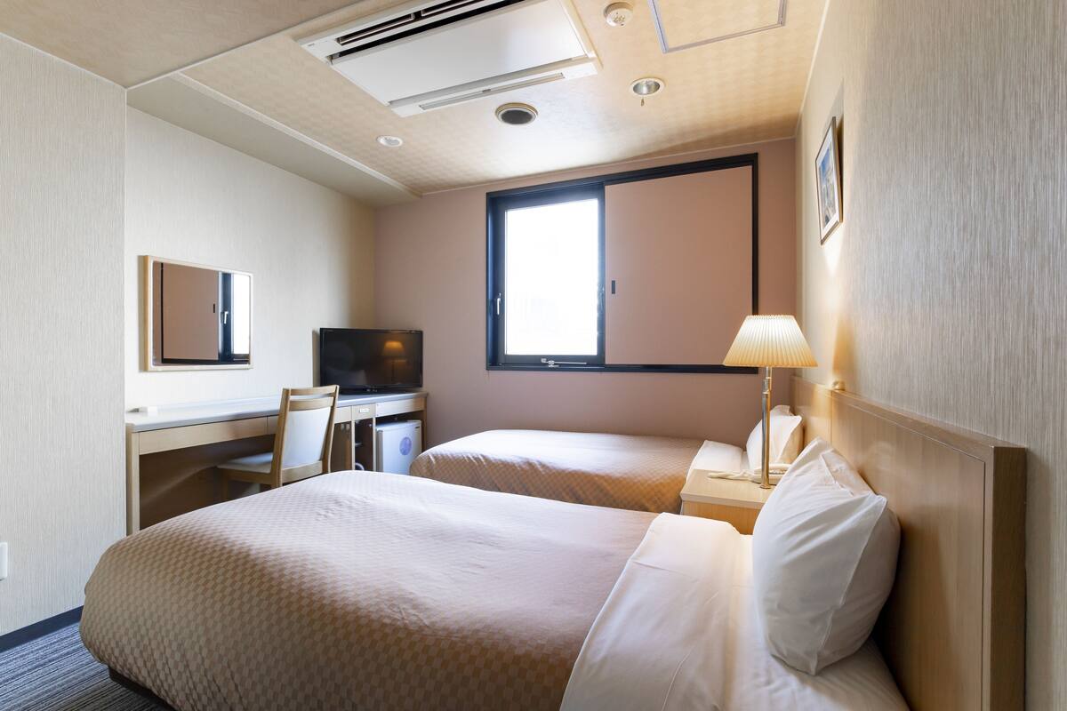 A hotel room with two beds, a desk with a chair, a television, and a window providing natural light.