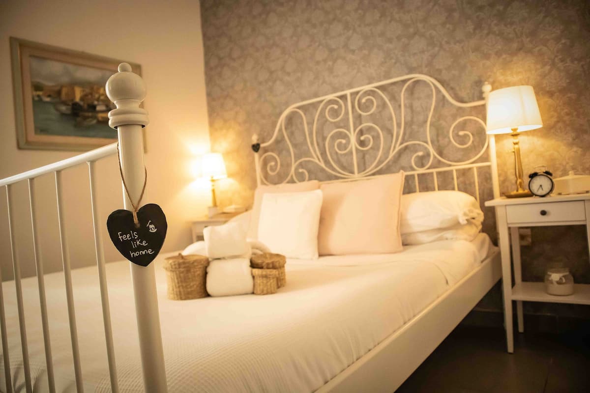 Una cama bien hecha con ropa blanca, un cabecero decorativo y una mesita de noche con lámpara y despertador.