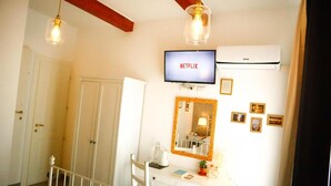 Une pièce avec une commode blanche, un miroir doté d’un cadre décoratif, un téléviseur affichant le logo « NETFLIX » et un ventilateur de plafond.