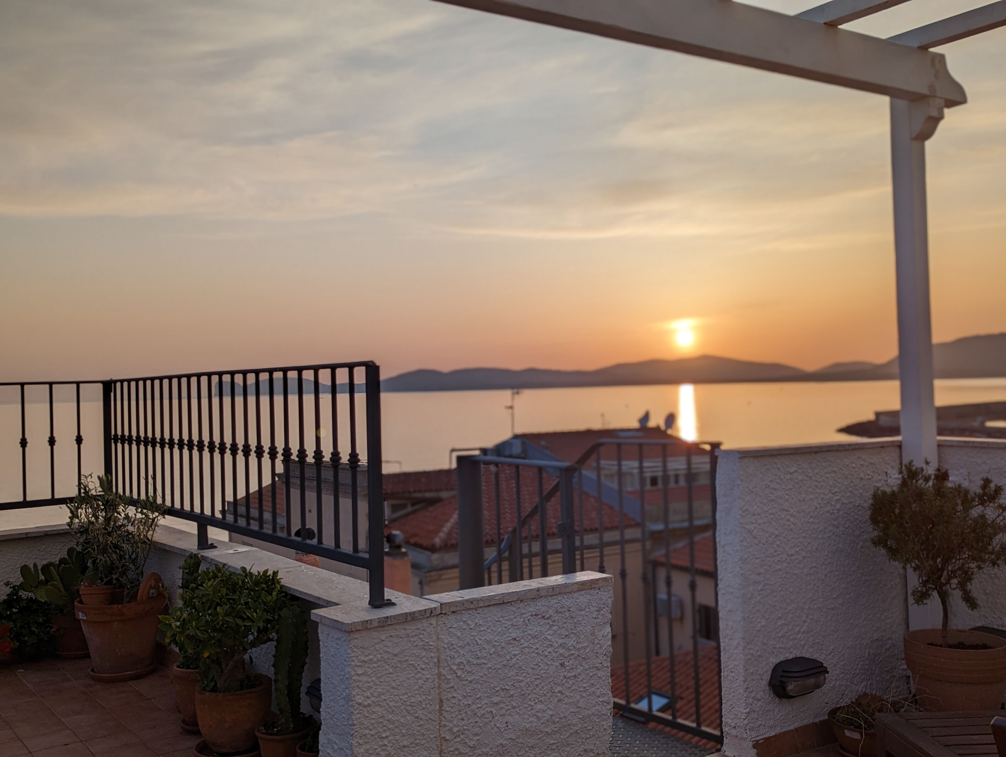 Un balcone con ringhiera, piante in vaso e vista sul tramonto sull'acqua.