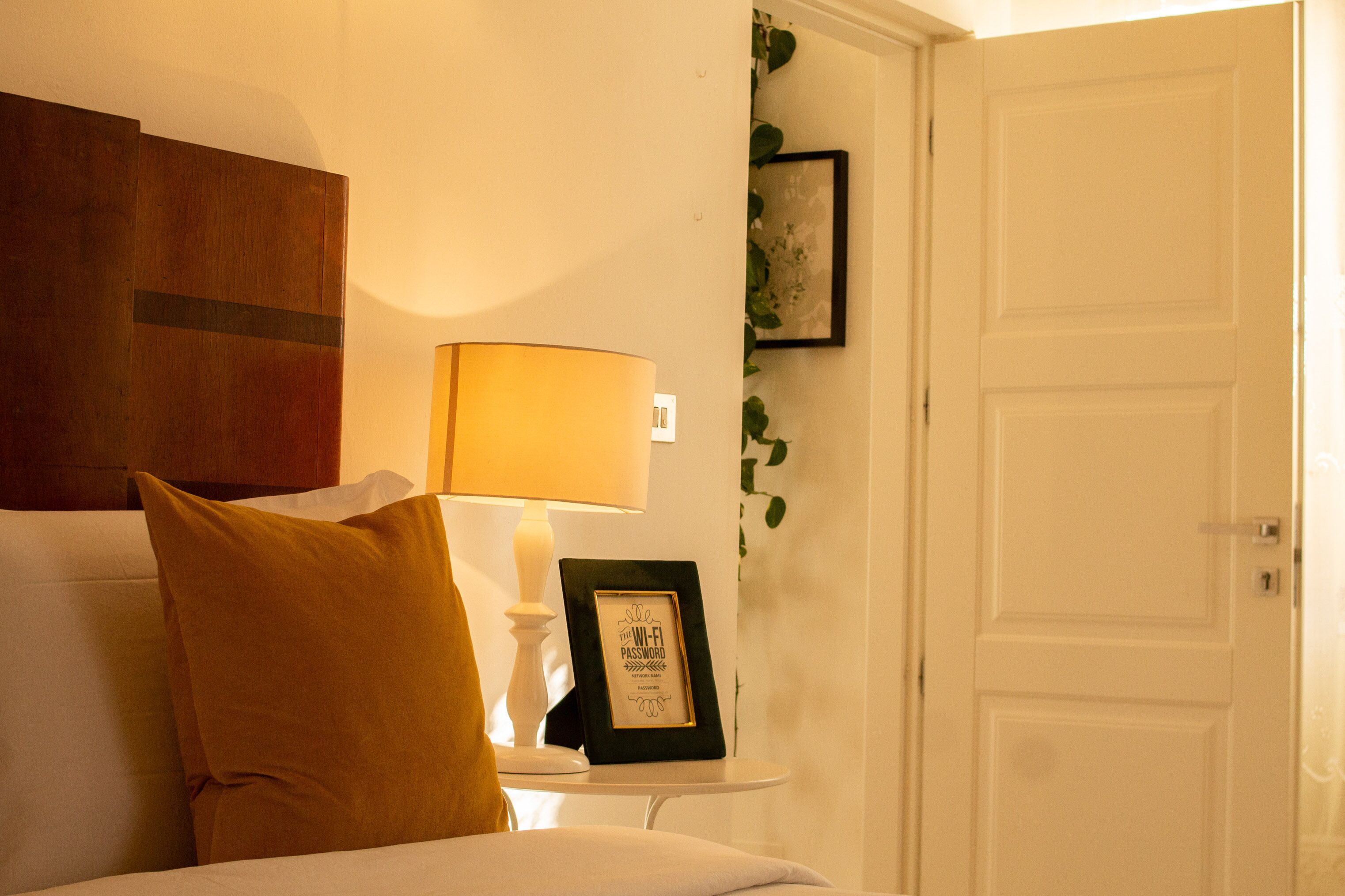 A neatly arranged bedroom with a bed, a bedside table with a lamp, a framed picture, and a door.