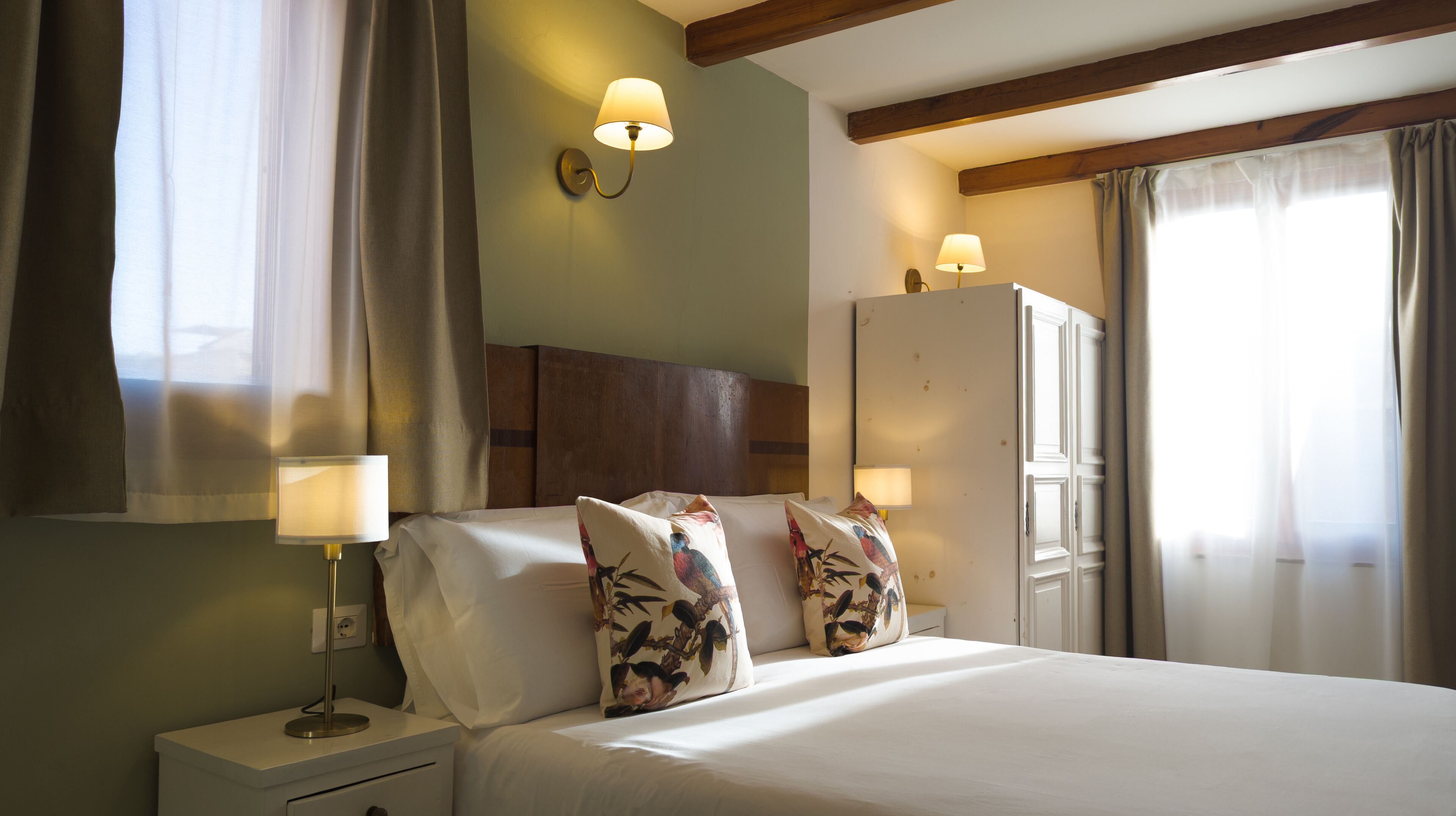 A hotel room with a bed, two bedside lamps, a nightstand, a wardrobe, and a window with curtains.