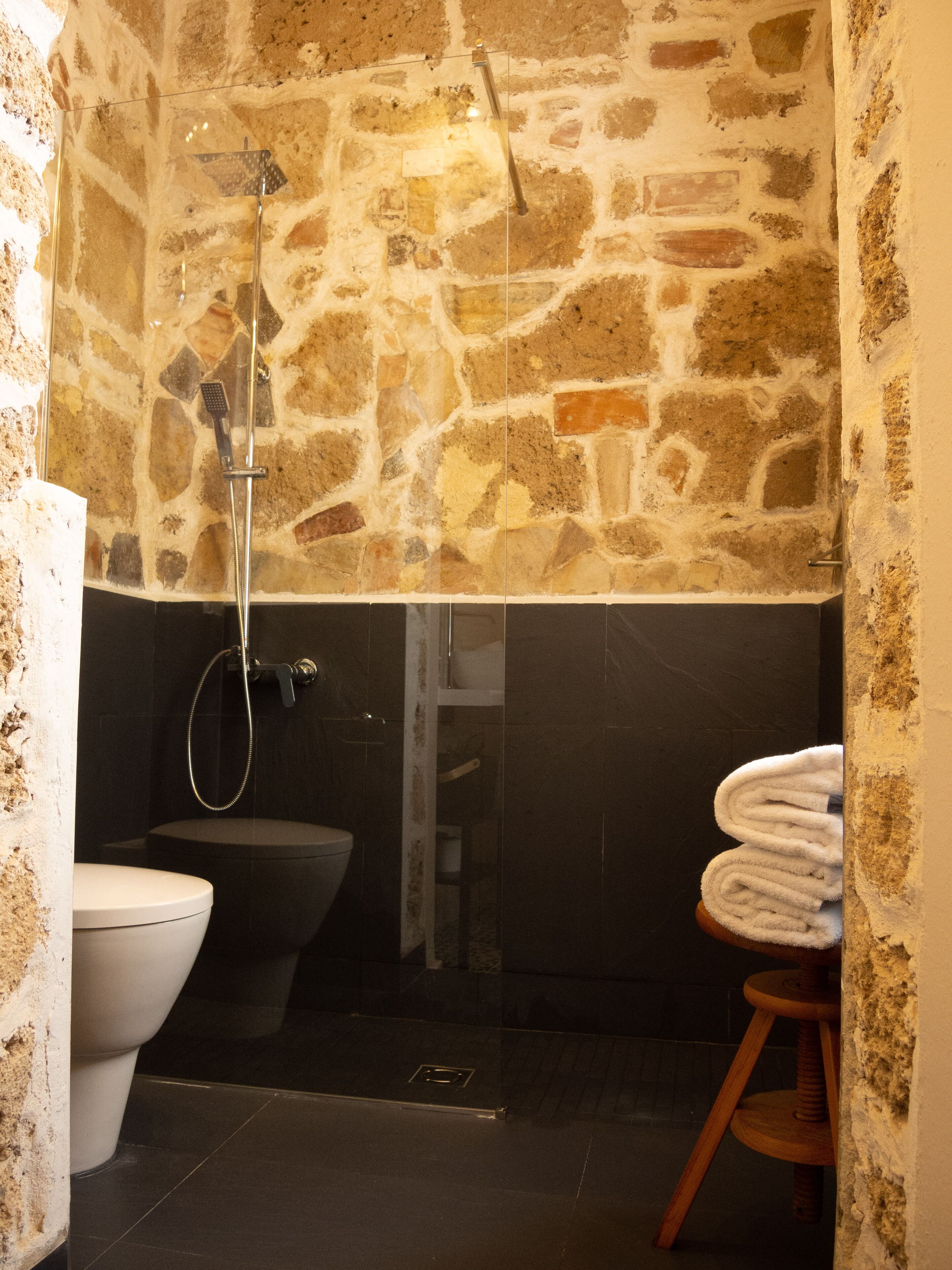 Un baño con pared de piedra, inodoro, ducha y taburete de madera con una toalla.