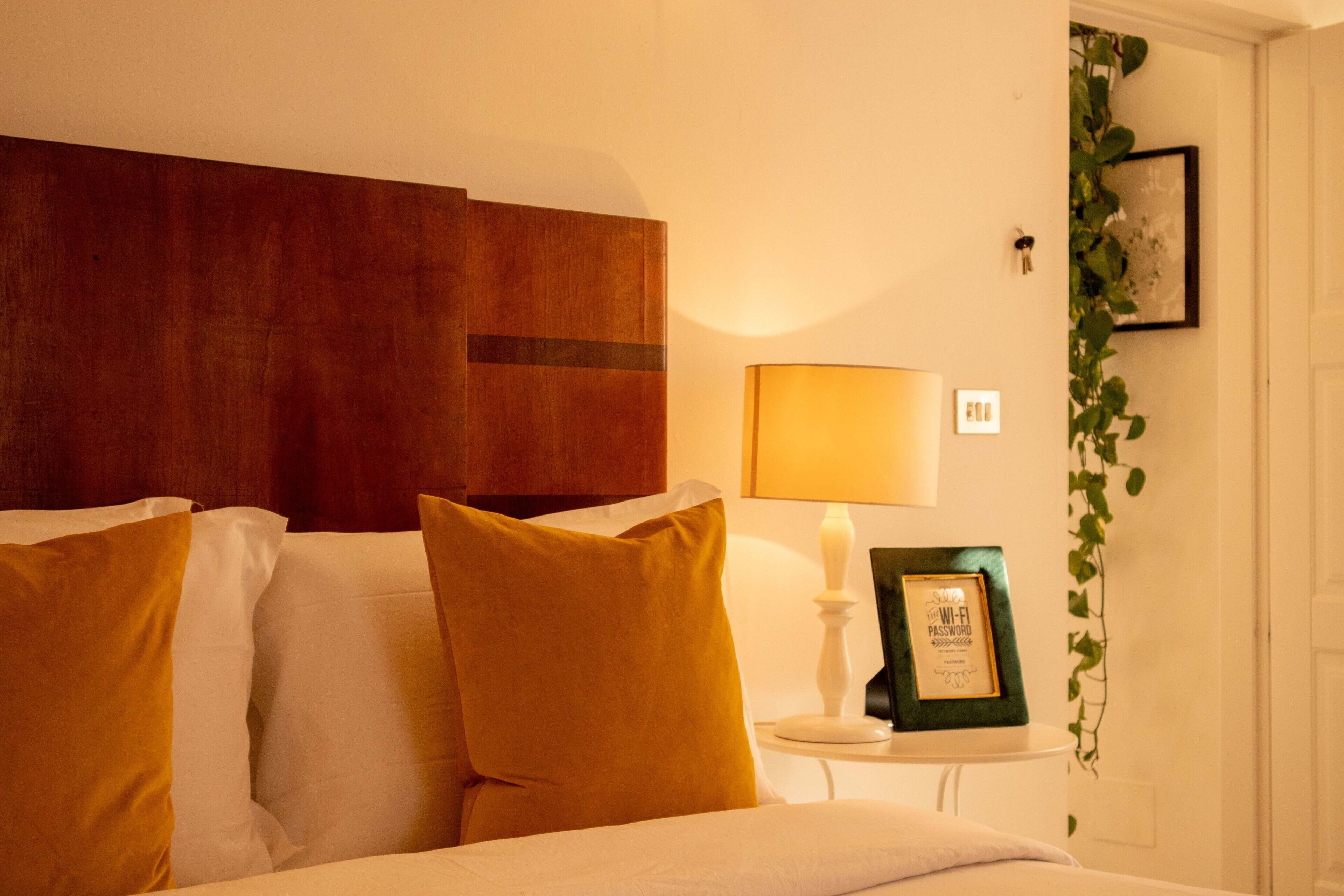 A hotel room with a wooden headboard, a bedside table with a lamp, a framed picture, and a potted plant.
