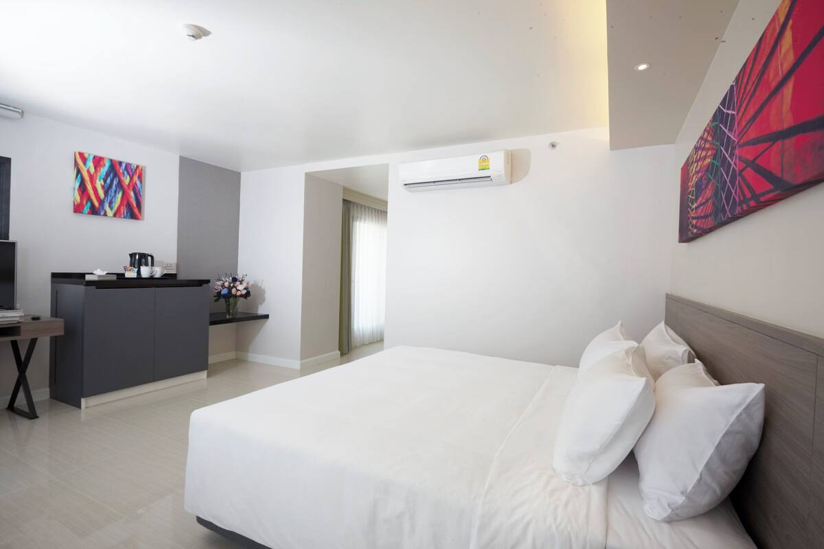 A hotel room with a large bed, a desk with a coffee maker, a colorful abstract painting, and an air conditioning unit.