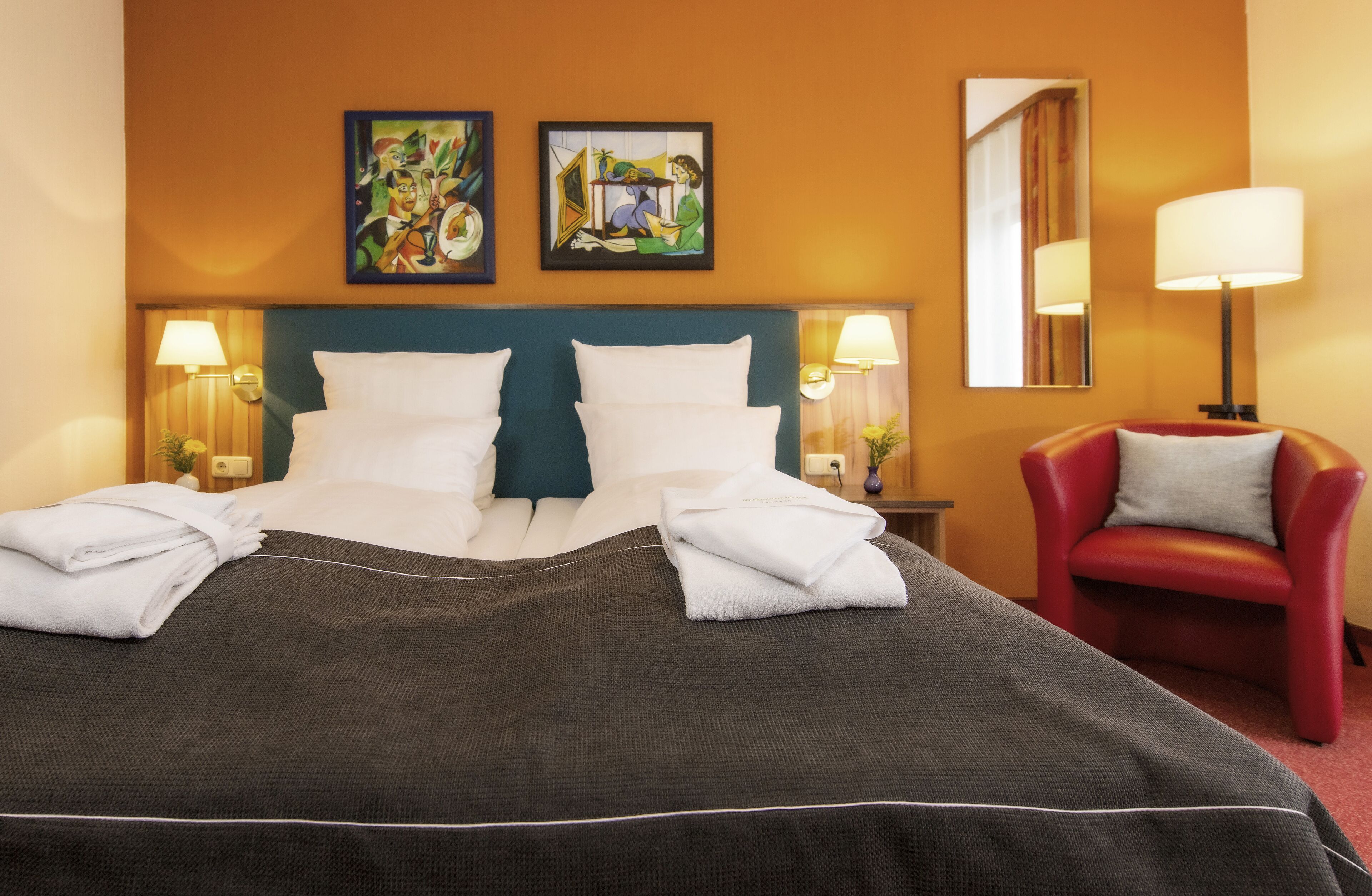 Een hotelkamer met een groot bed, twee schilderijen aan de muur, een rode stoel en twee bedlampen.