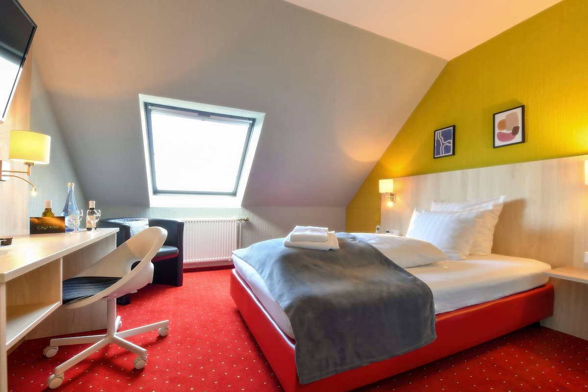 Een hotelkamer met een rood tapijt, een bed met witte lakens, een bureau met een stoel, een raam en een kleine tafel met een lamp.