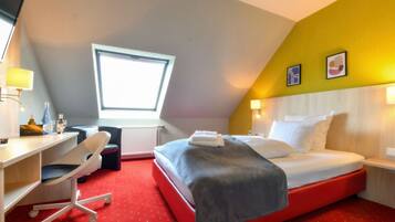 Een hotelkamer met een rood tapijt, een bed met witte lakens, een bureau met een stoel, een raam en een kleine tafel met een lamp.