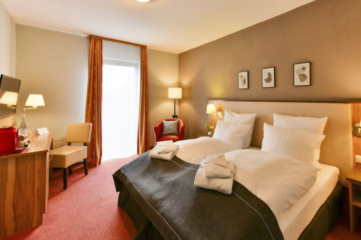 Een hotelkamer met een groot bed, twee nachtkastjes met lampen, een bureau met telefoon, een stoel en een raam met gordijnen.
