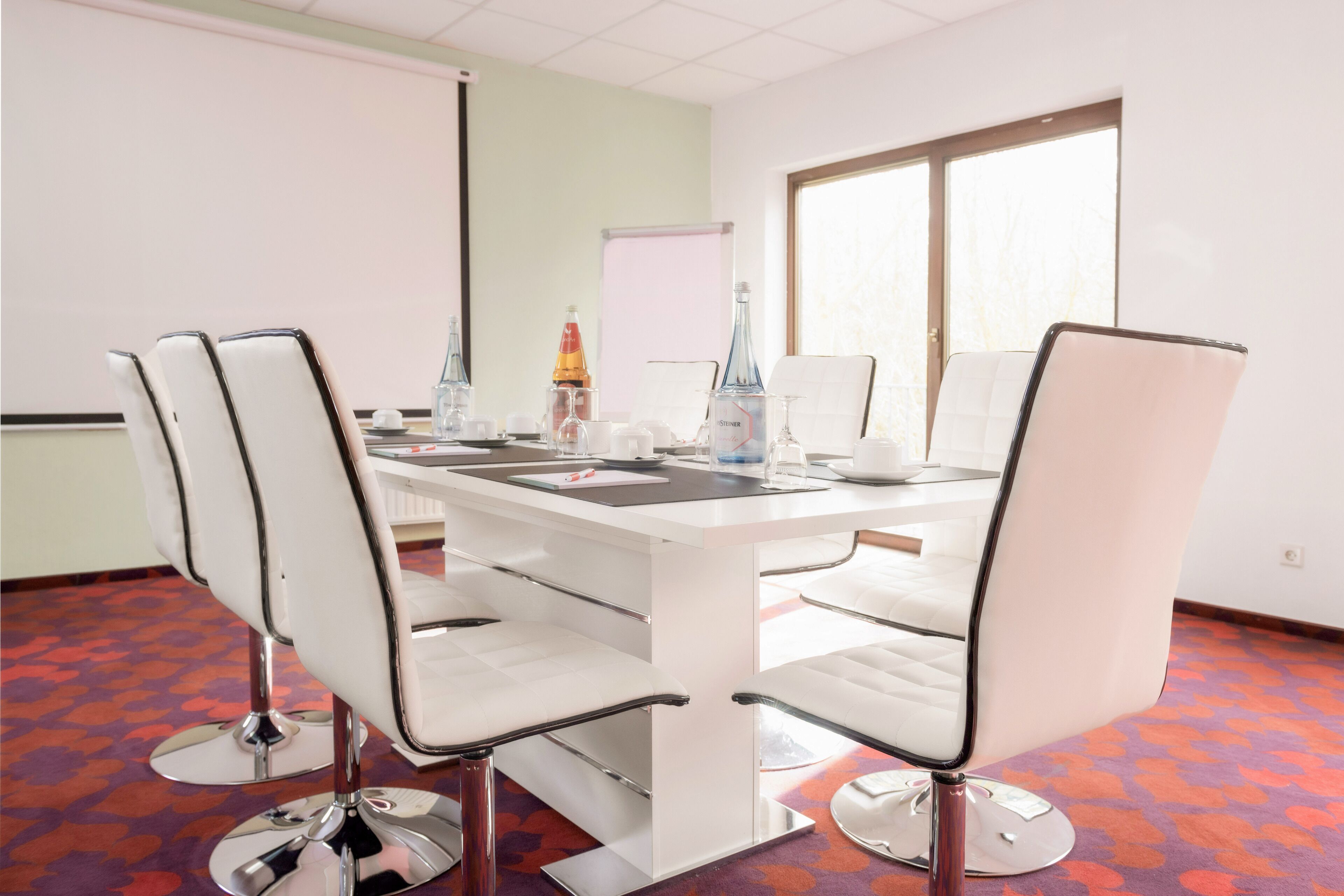 Een vergaderruimte met een witte tafel, vier witte stoelen, een projectiescherm en een raam met jaloezieën.