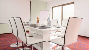 Un salón de conferencias con una mesa blanca, cuatro sillas blancas, una pantalla de proyector y una ventana con persianas.