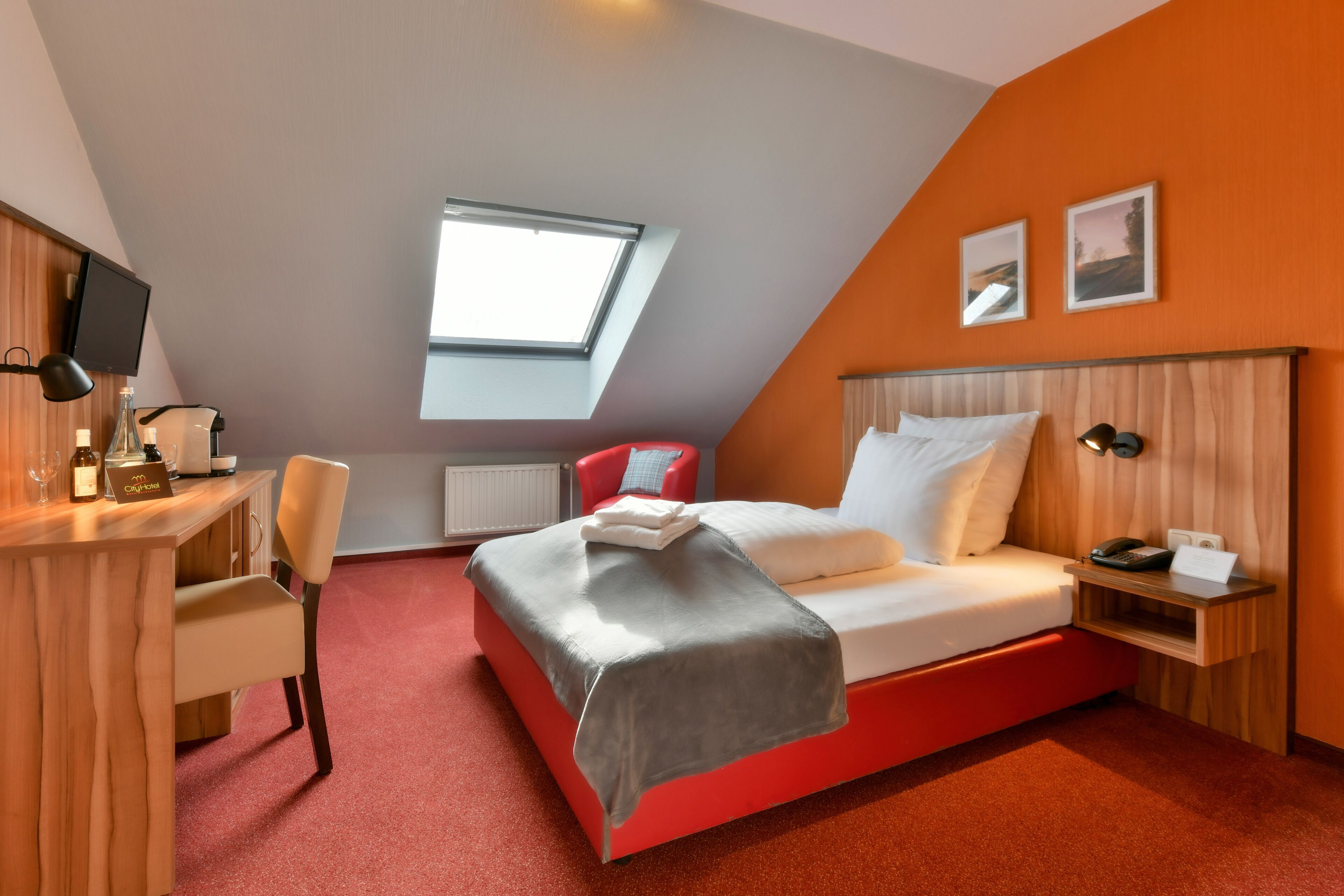 Ein modernes Hotelzimmer mit schrägverdachter Decke, einem Oberlicht, einem Bett mit rotem Gestell, einem Schreibtisch mit Stuhl und einem kleinen Nachttisch mit Lampe.