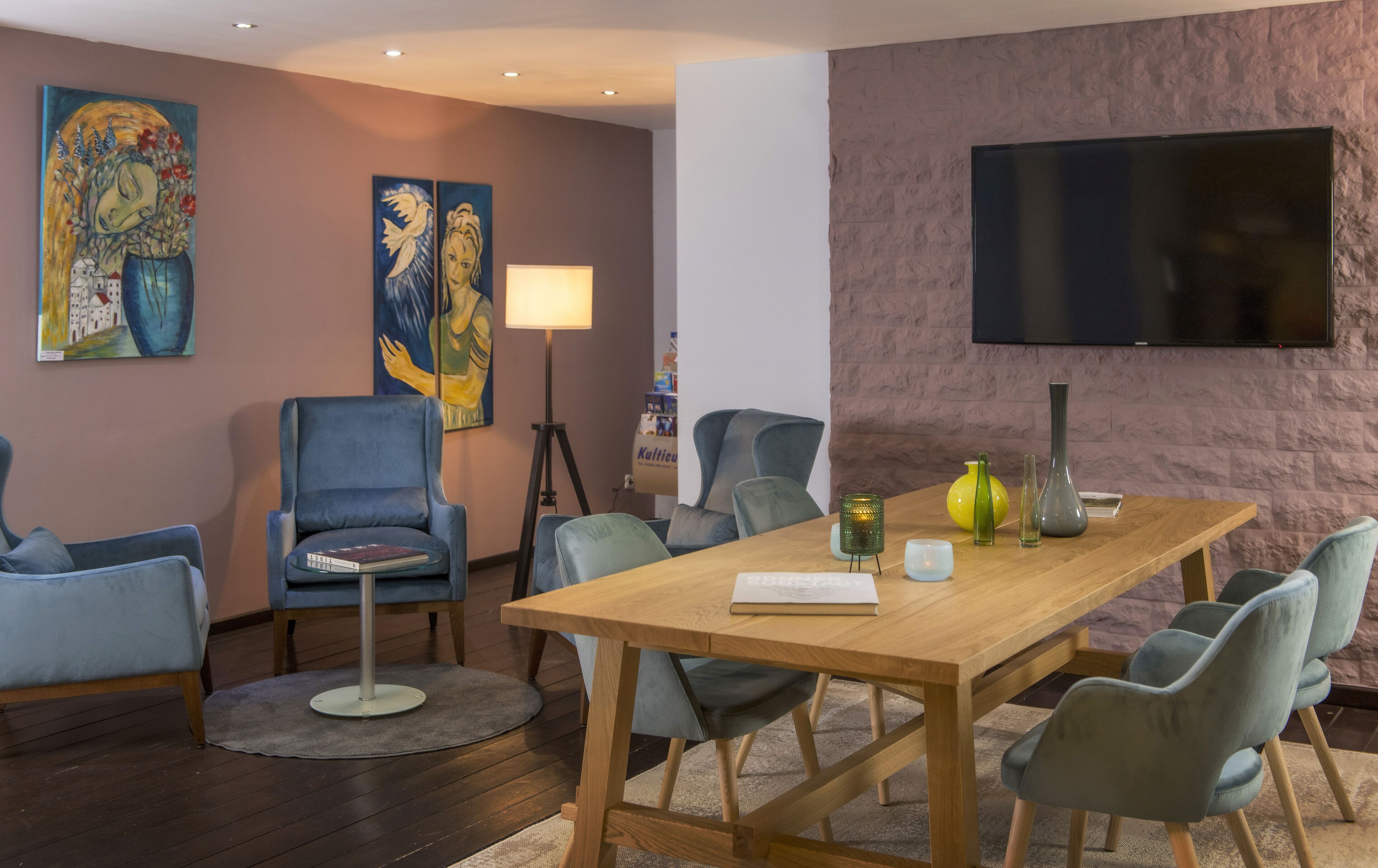 Een moderne vergaderruimte met een houten tafel, blauwe stoelen, een staande lamp en kunstwerken aan de muren.