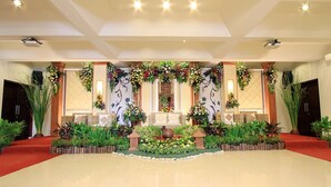 A spacious hall with a central stage, surrounded by lush greenery and decorative floral arrangements.