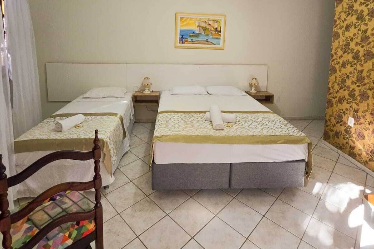 Quarto de hotel com duas camas, uma cadeira, um quadro na parede e piso ladrilhado.