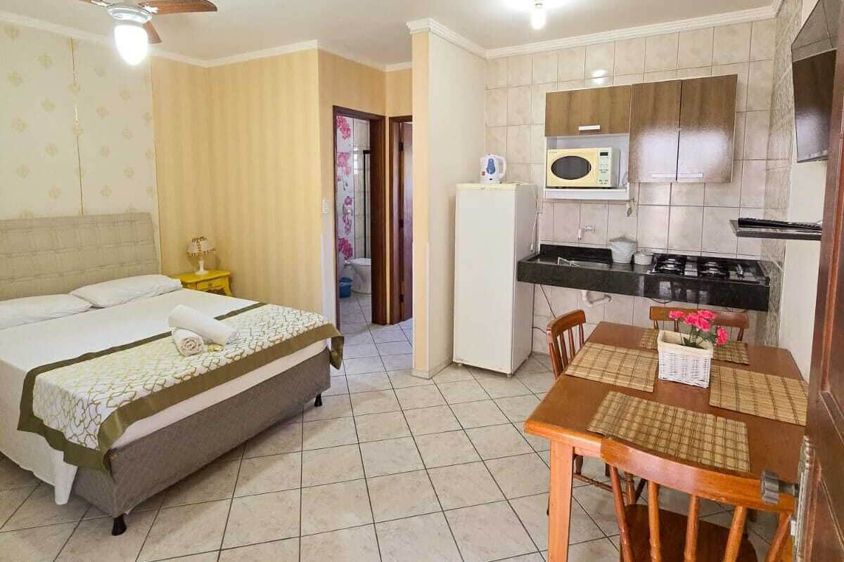 Quarto de hotel compacto com cama, pequeno espaço de cozinha, mesa de jantar com cadeiras e ventoinha no teto.