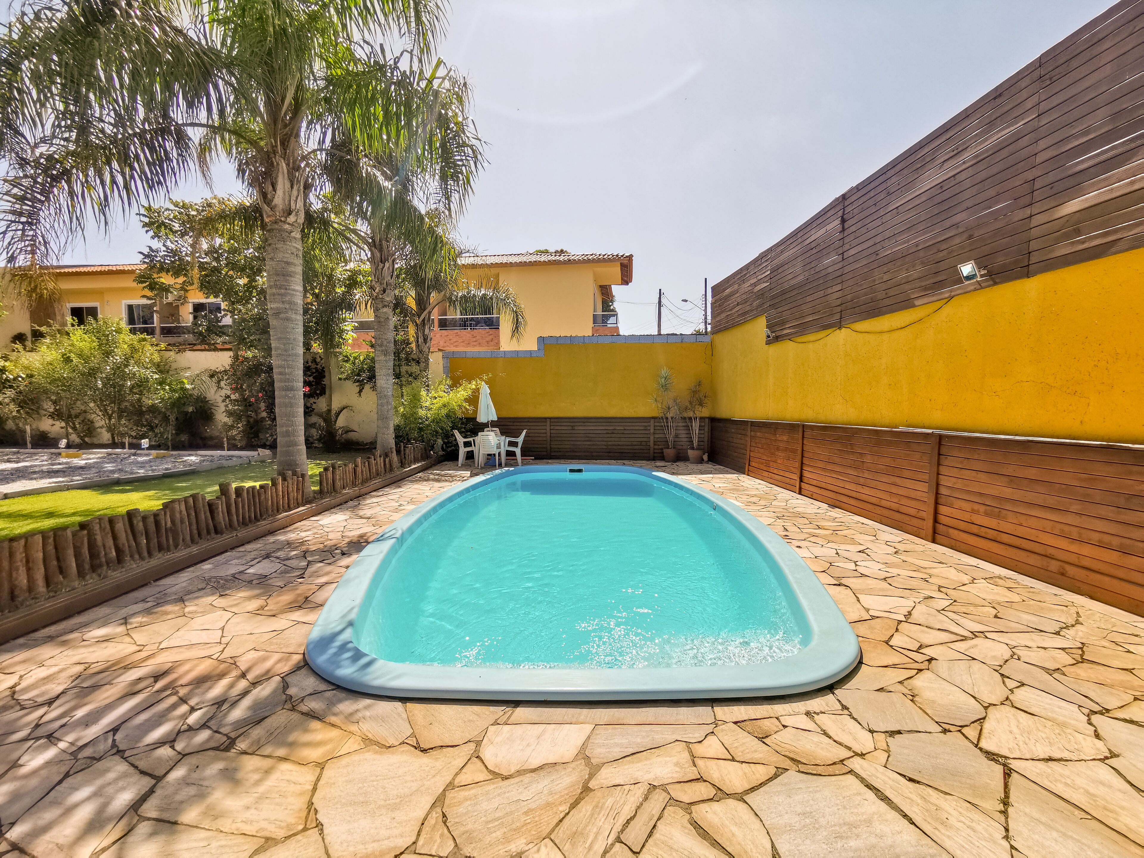 A rectangular swimming pool with a stone-tiled deck, surrounded by a wooden fence and a yellow wall.