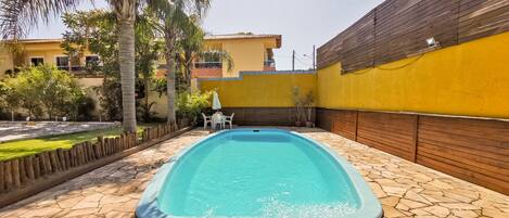 A rectangular swimming pool with a stone-tiled deck, surrounded by a wooden fence and a yellow wall.