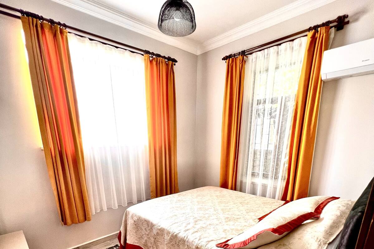 A hotel room with a bed, a red patterned carpet, and two windows with orange curtains.