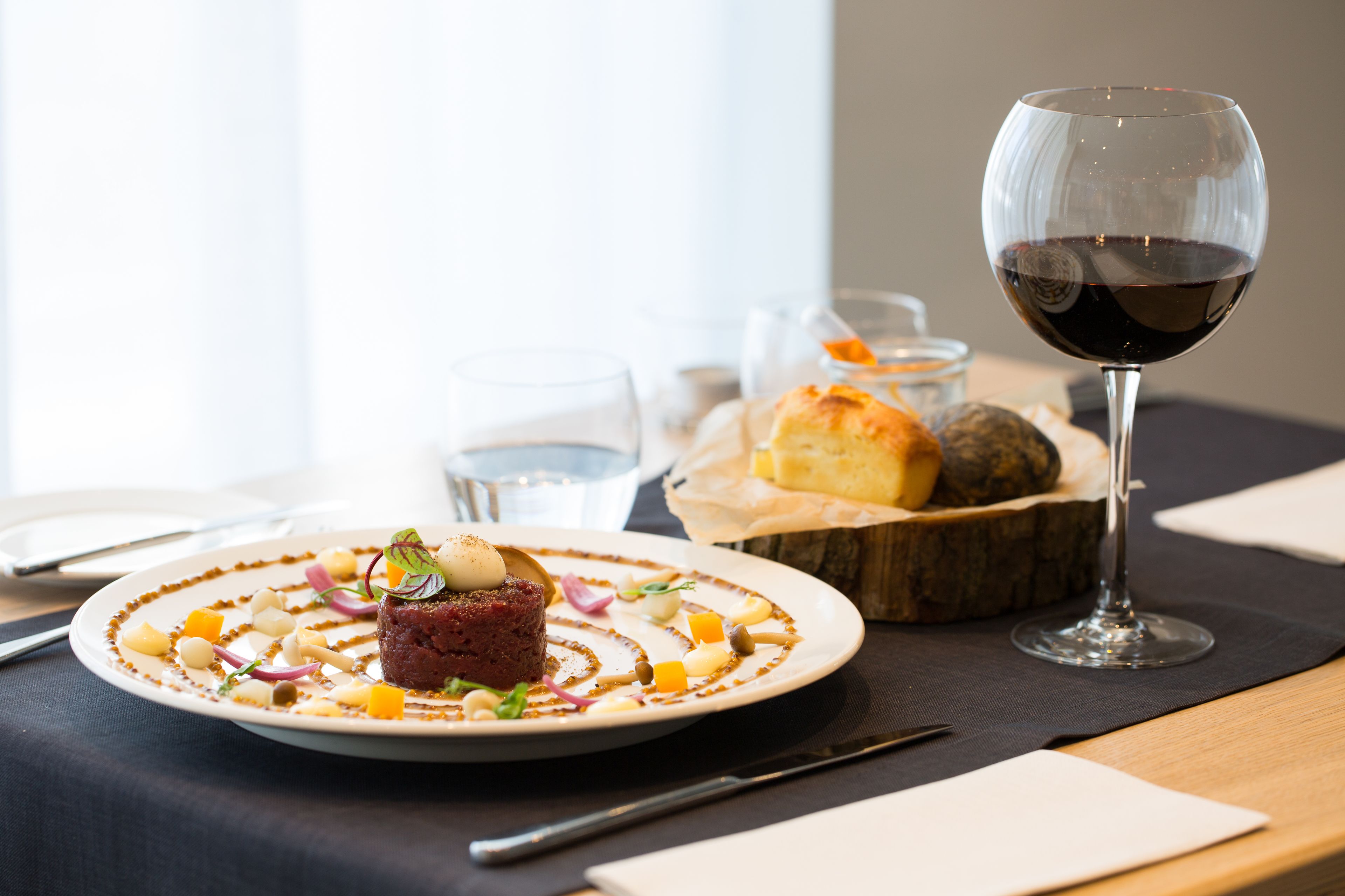 A dining table with a plate of food, a glass of red wine, and a basket of bread.