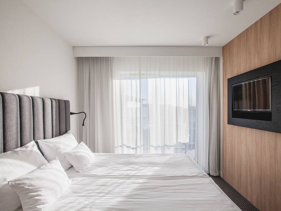 A modern hotel room with a large bed, a mounted TV, and a view of the city through sheer curtains.