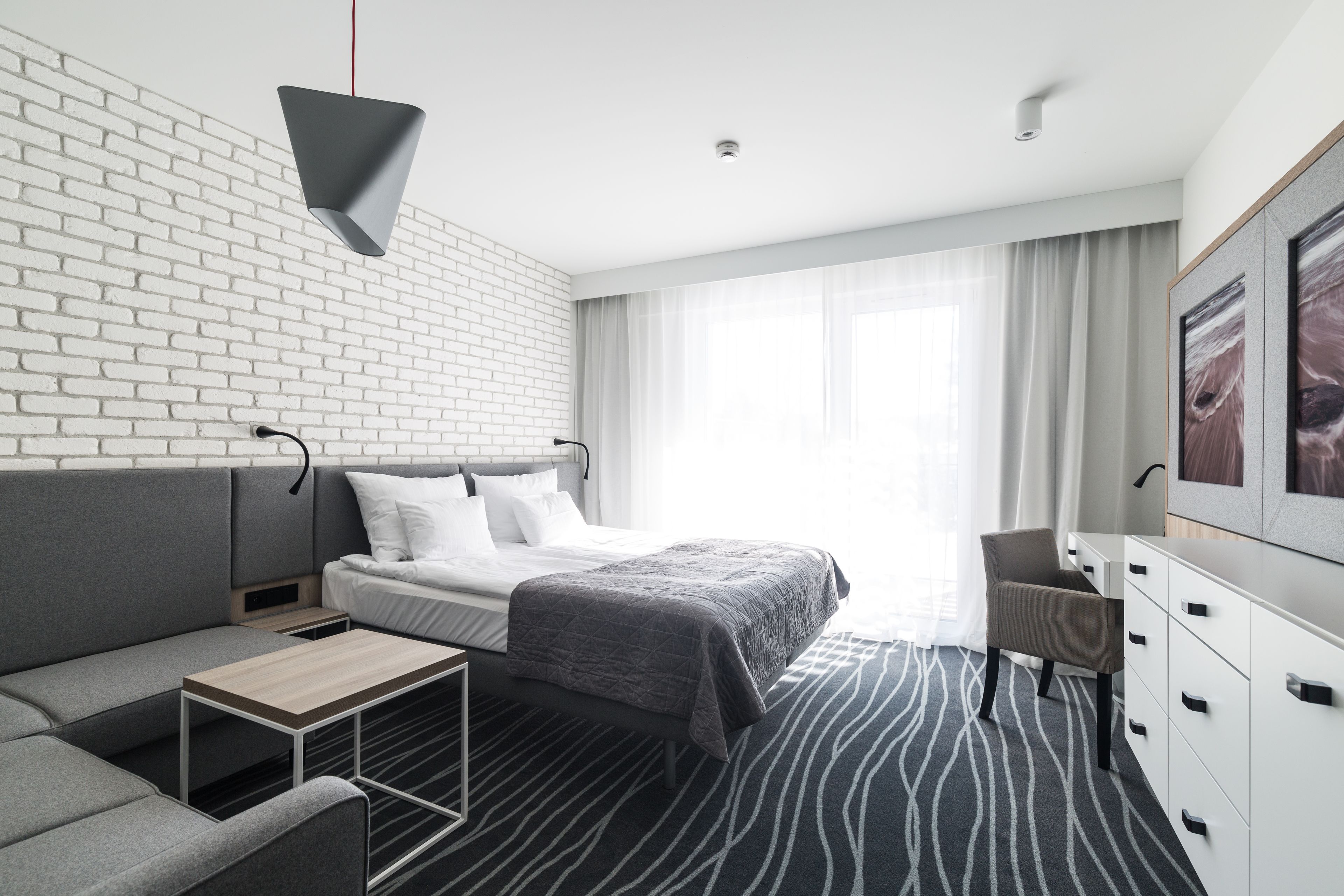 Ein modernes Hotelzimmer mit Backsteinwand, einem Bett mit grauer Decke, einem grauen Sofa, einem kleinen Tisch, einem Schreibtisch und einem Kleiderschrank.