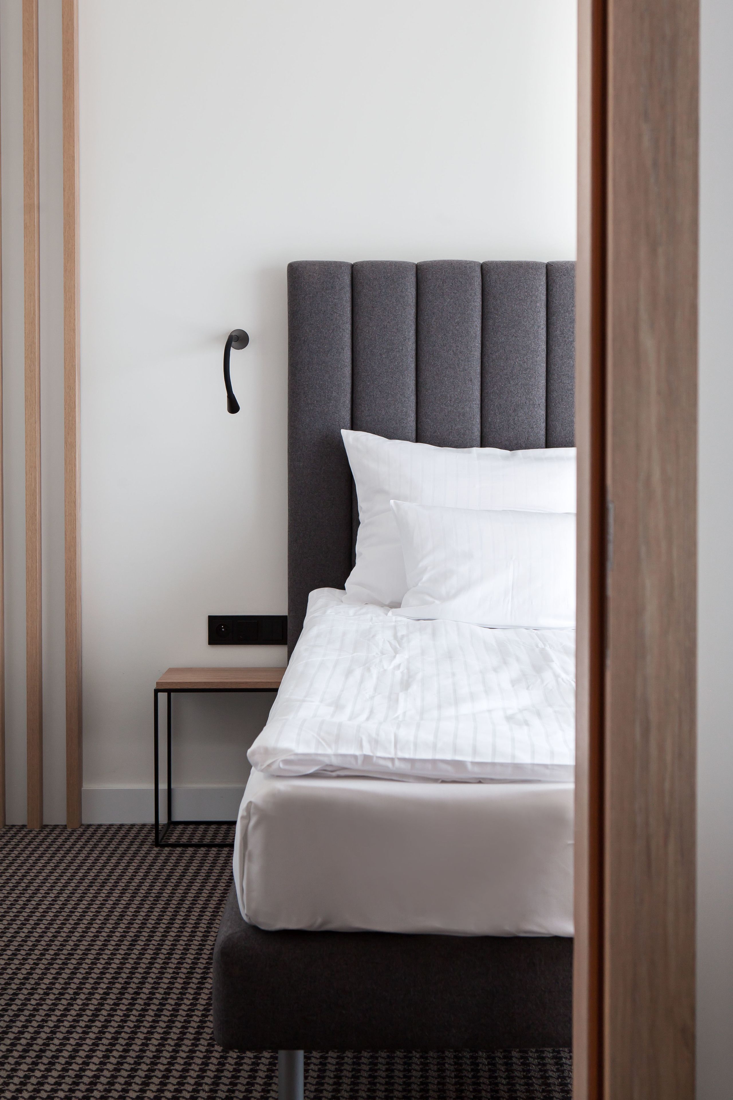 A neatly made bed with white linens, a grey upholstered headboard, and a small bedside table with a black lamp.