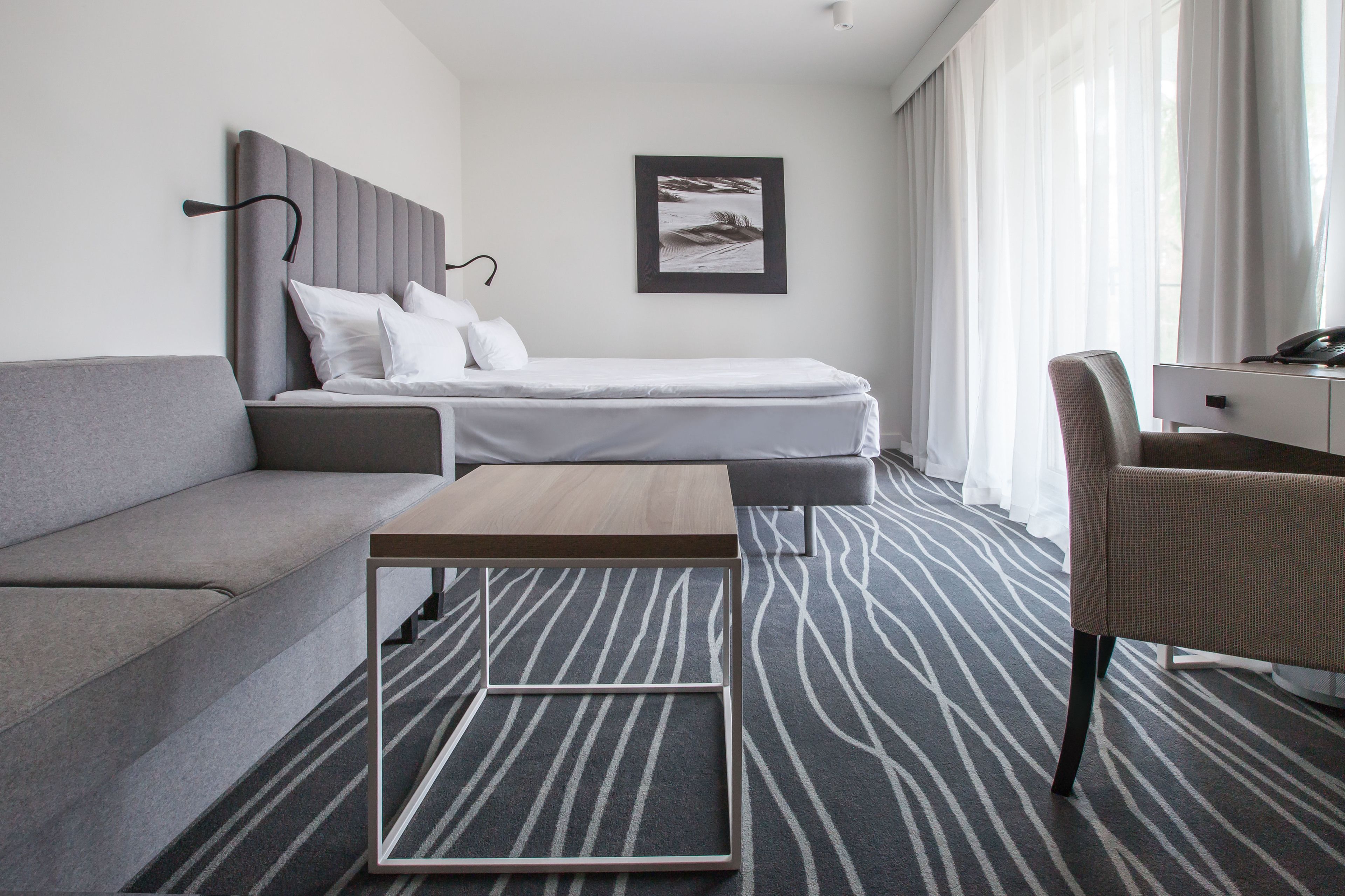 A modern hotel room with a grey sofa, a single bed, a small table, and a chair.