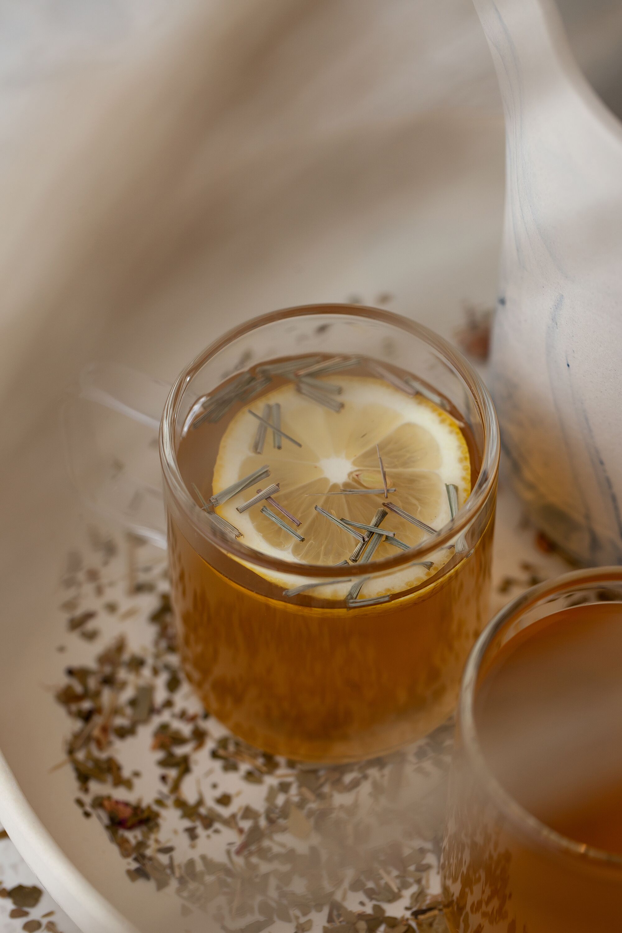 A glass of tea with a slice of lemon and a tea infuser, placed on a tray with loose tea leaves.