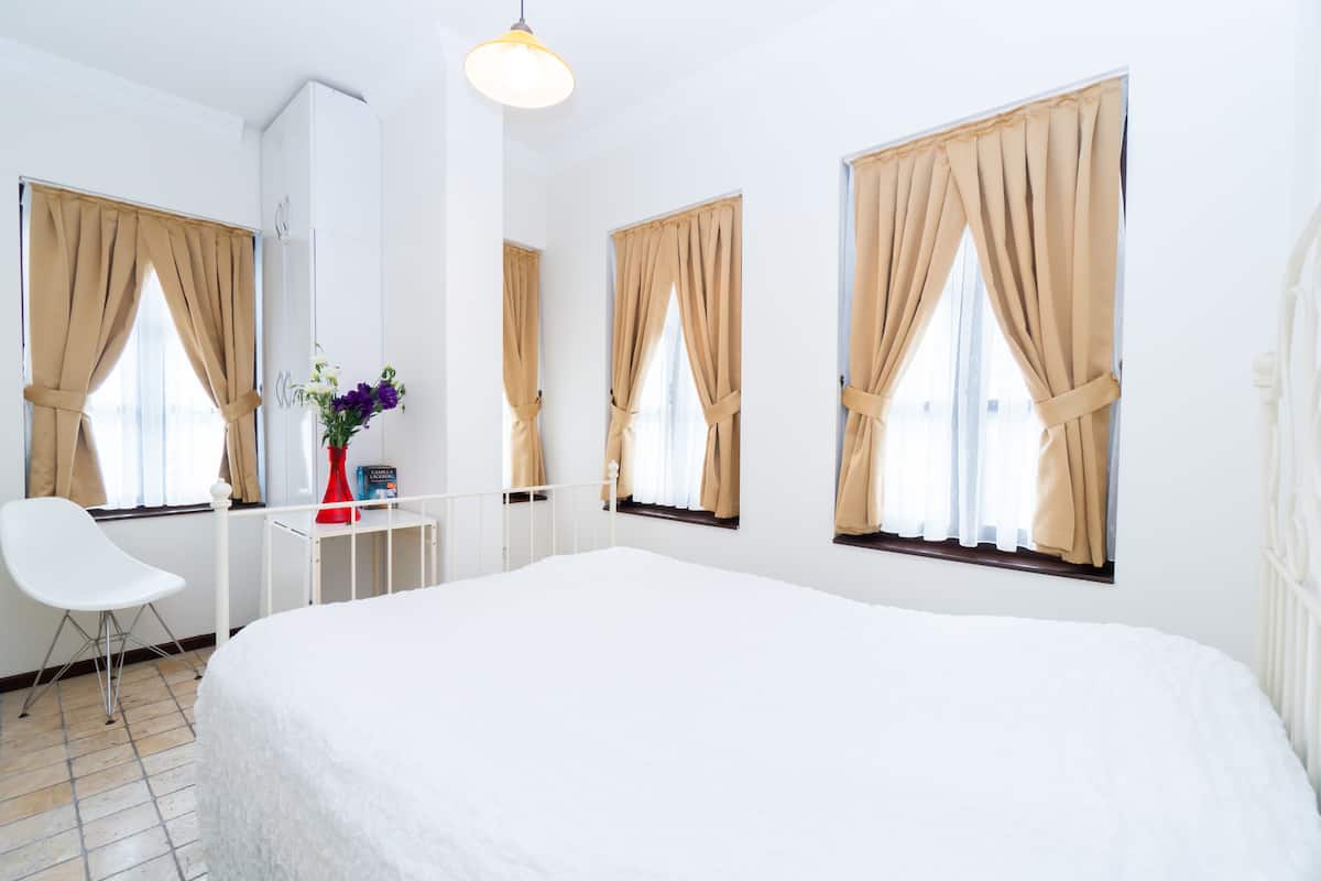 Habitación de hotel con una cama grande, una silla blanca, un escritorio con un florero y cortinas color beige en las ventanas.