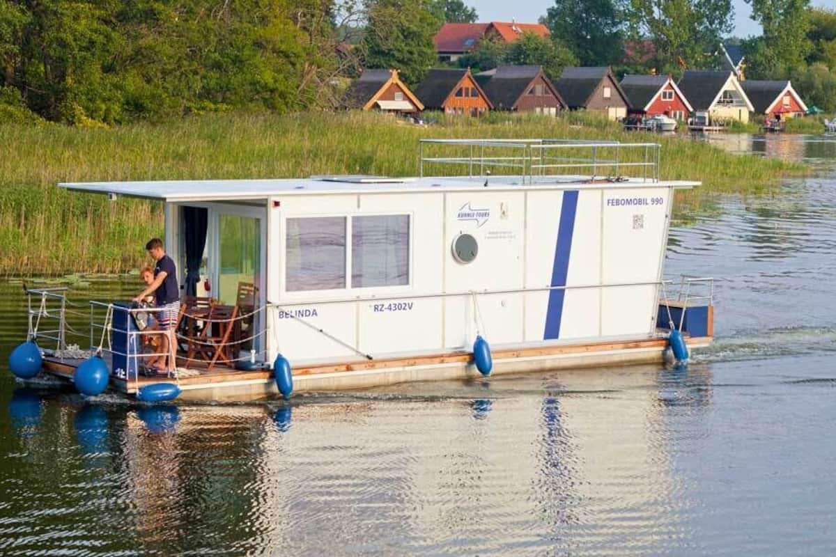Ein Hausboot namens 'BELINDA' mit einer kleinen Deckfläche und einem überdachten Bereich, das auf ruhigem Gewässer schwimmt.