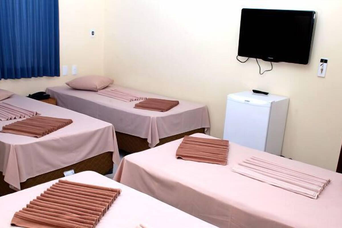 Une chambre dâhĂŽtel avec trois lits, chacun recouvert dâune couverture rose et agrĂ©mentĂ© dâune serviette marron. Un tĂ©lĂ©viseur est fixĂ© au mur, et on aperçoit un petit appareil Ă©lectromĂ©nager blanc.