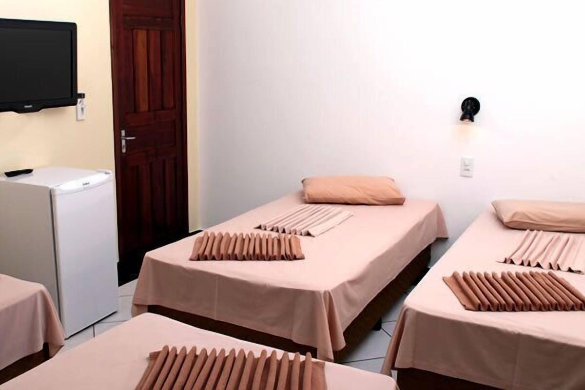 Une chambre avec trois lits, chacun recouvert dâune couvre-lit rose et agrĂ©mentĂ© dâun coussin au motif unique. Un tĂ©lĂ©viseur Ă Ă©cran plat est fixĂ© au mur et un rĂ©frigĂ©rateur se trouve dans un coin de la piĂšce.