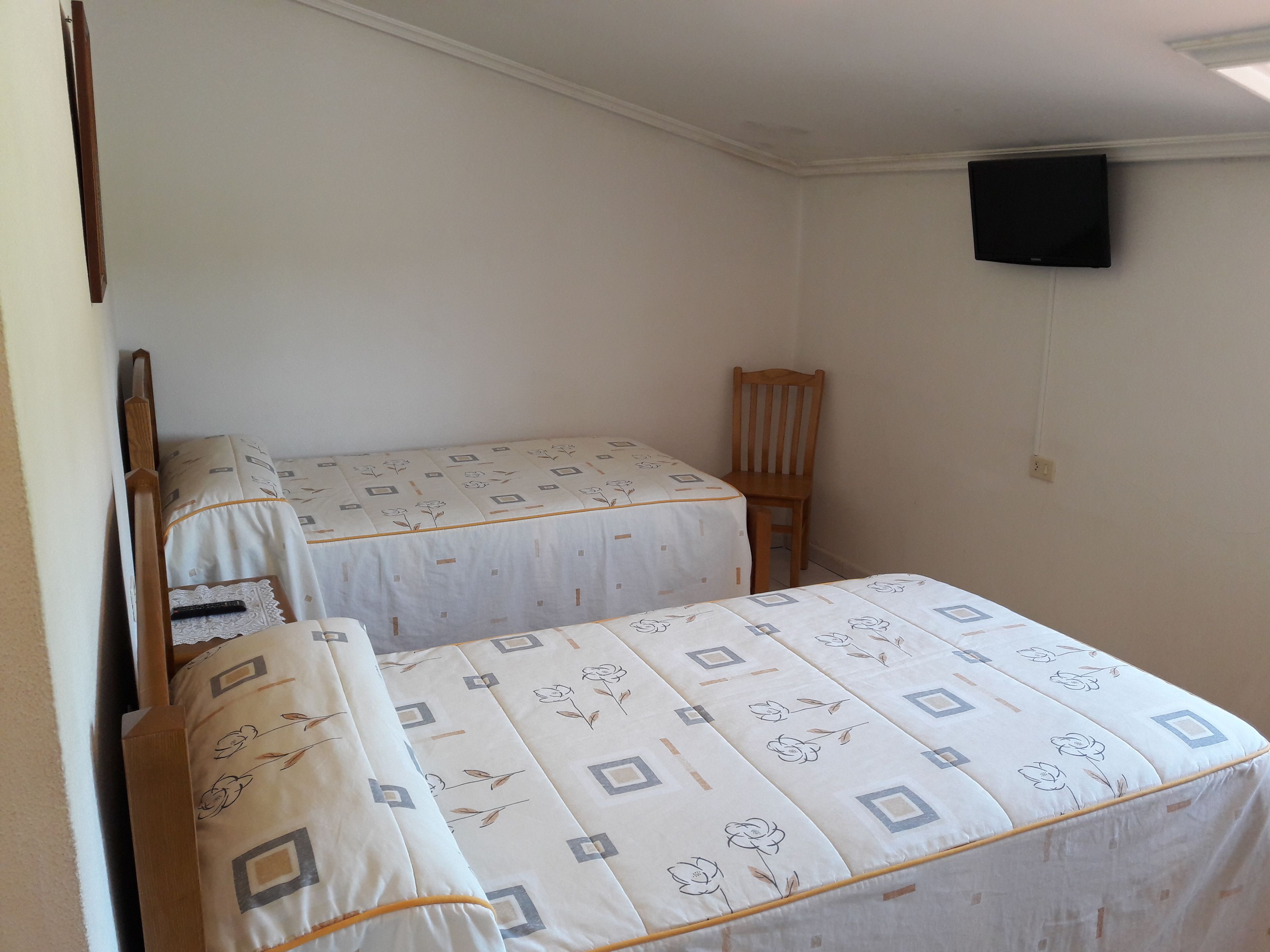 A small room with two beds, a wooden chair, and a television mounted on the wall.