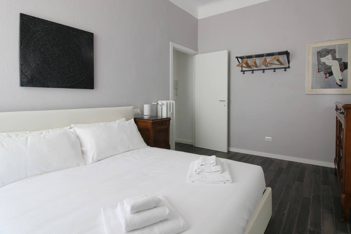 A hotel room with a neatly made bed, a door leading to another room, a coat rack with hangers, and a framed picture on the wall.