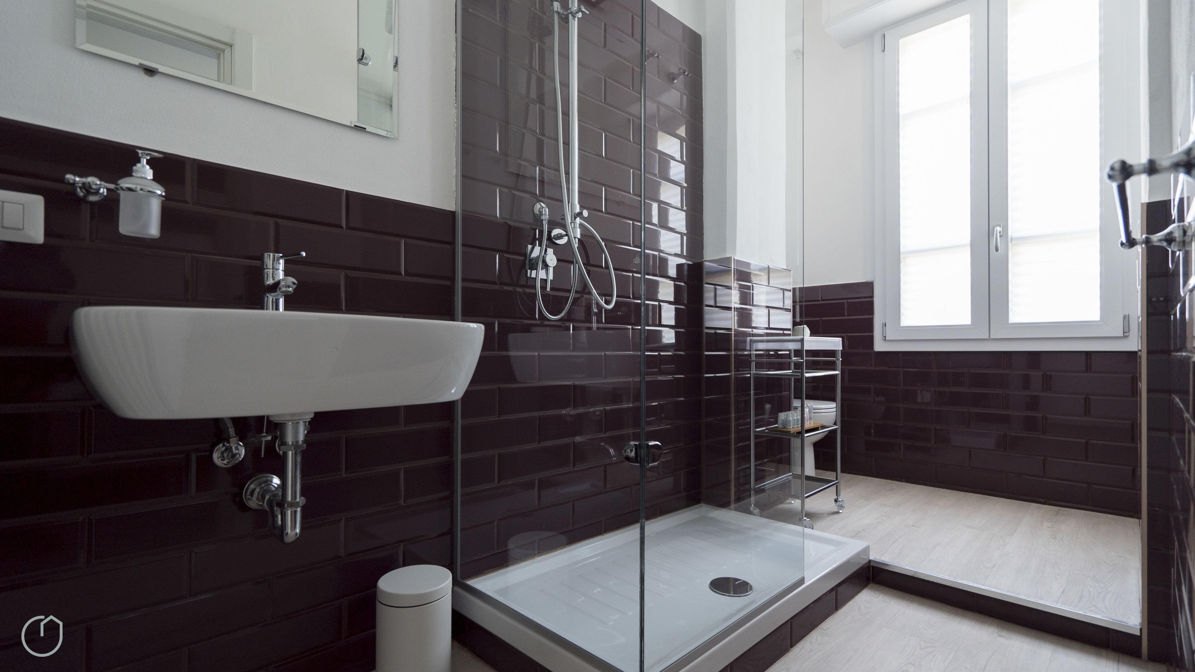A modern bathroom with a large white sink, dark tiled walls, a glass shower enclosure, and a window providing natural light.