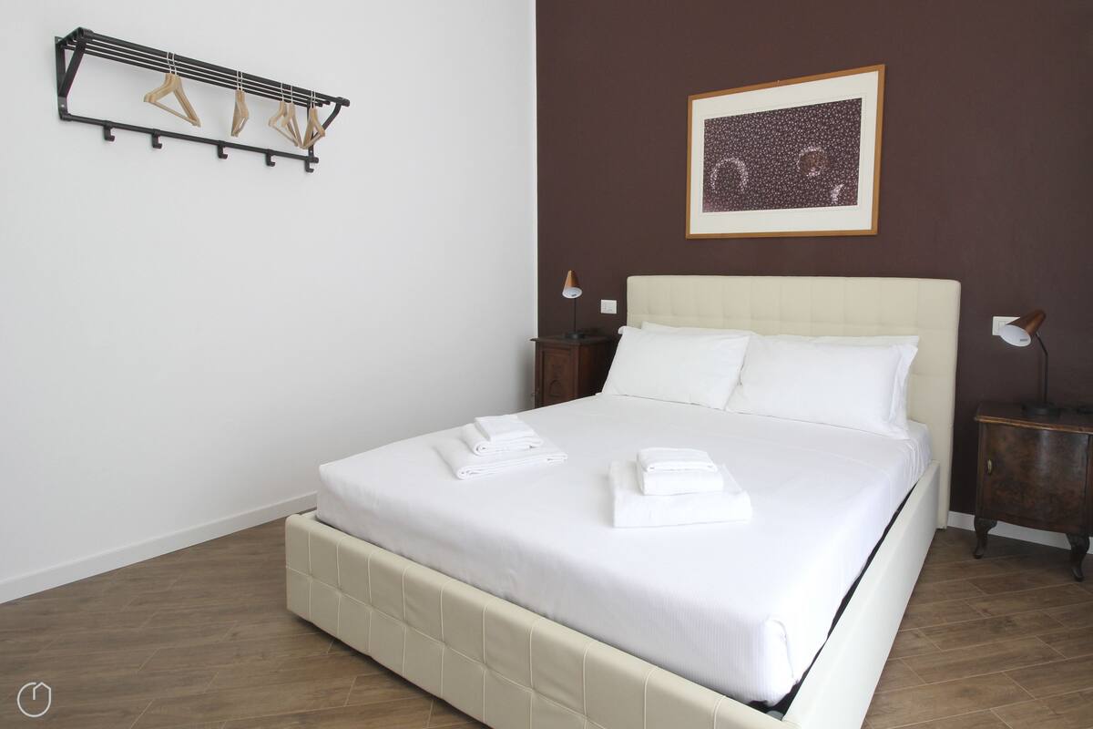 A neatly made bed with white linens and towels, a headboard, a framed picture on the wall, and a wooden floor.