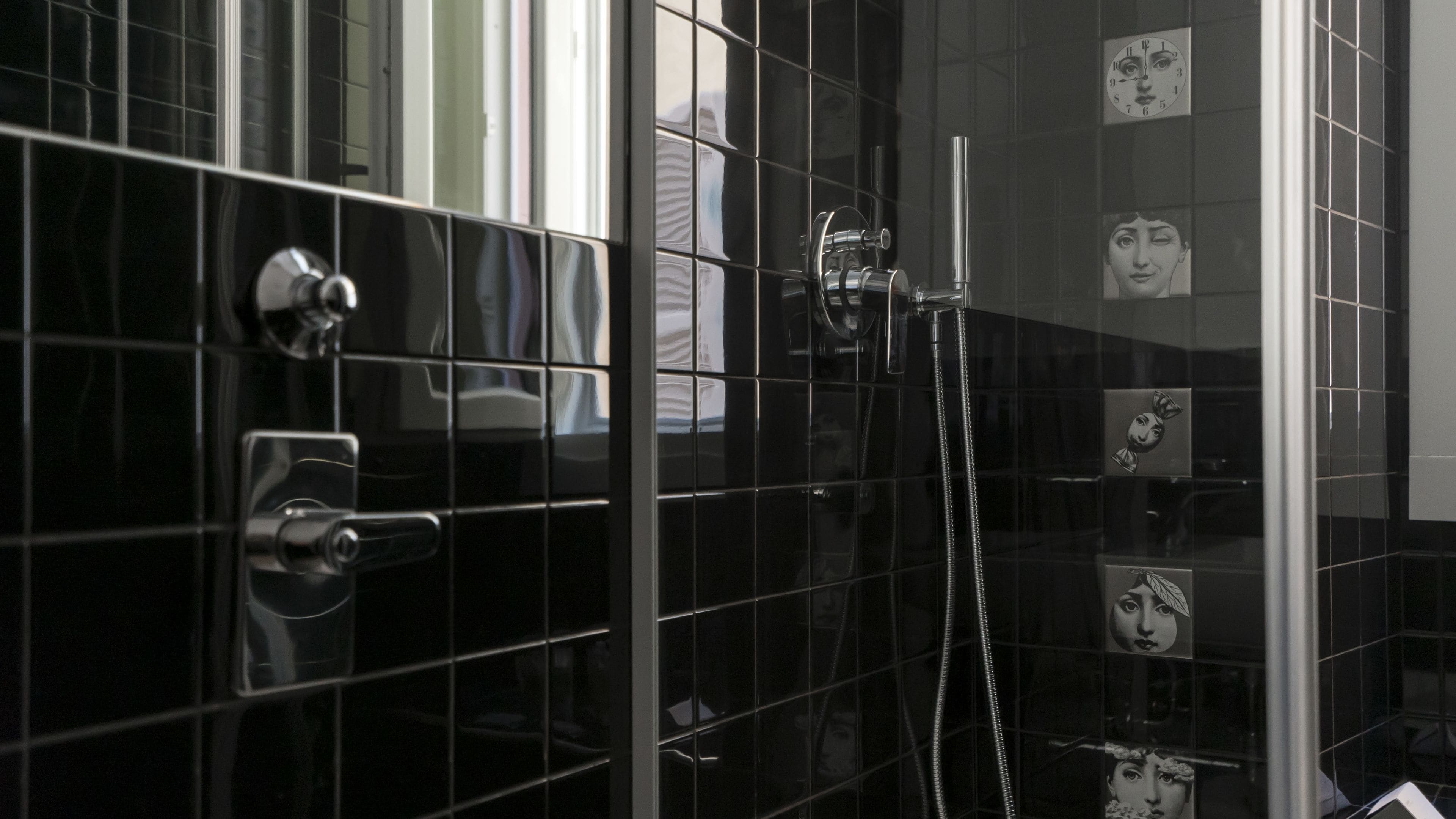 Une salle de bain avec des murs recouverts de carreaux noirs, une douche équipée d’un pommeau de douche amovible et un miroir qui reflète l’intérieur de la salle de bain.