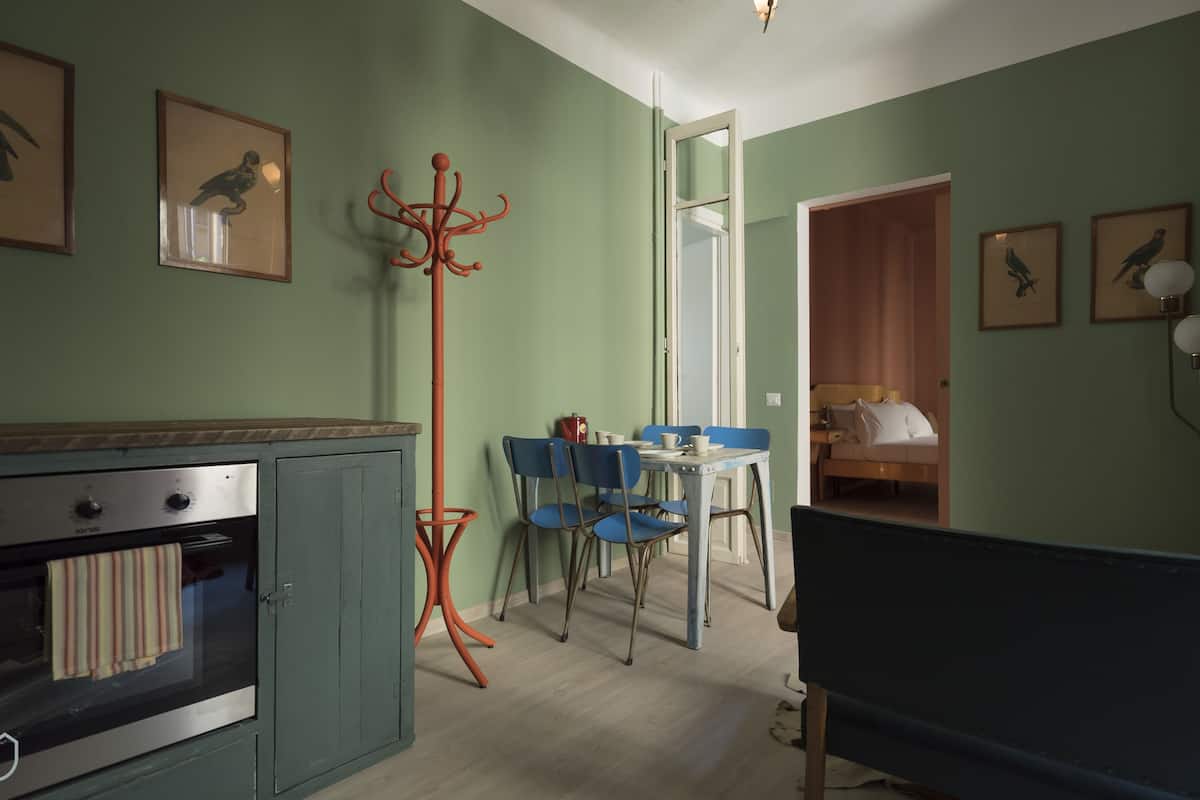 A room with a kitchenette, a dining area with blue chairs, a bed, and framed pictures on the walls.