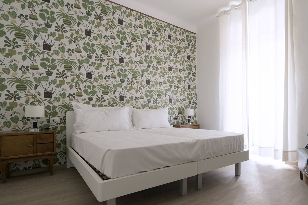 A bedroom with a bed, bedside tables, a nightstand, a chair, and a vase with flowers.