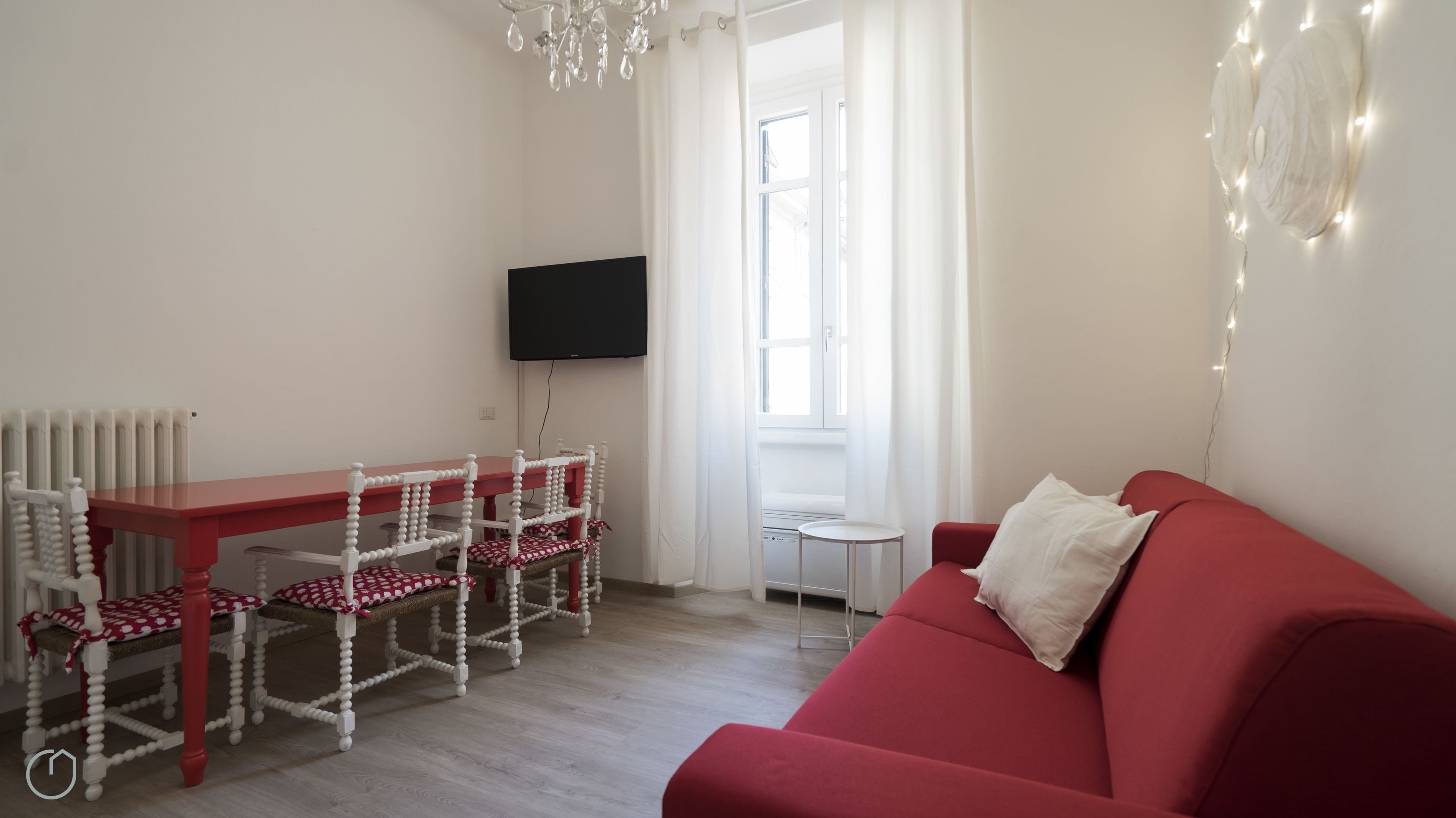 A living room with a red sofa, a wooden dining table with chairs, a TV mounted on the wall, and a radiator.