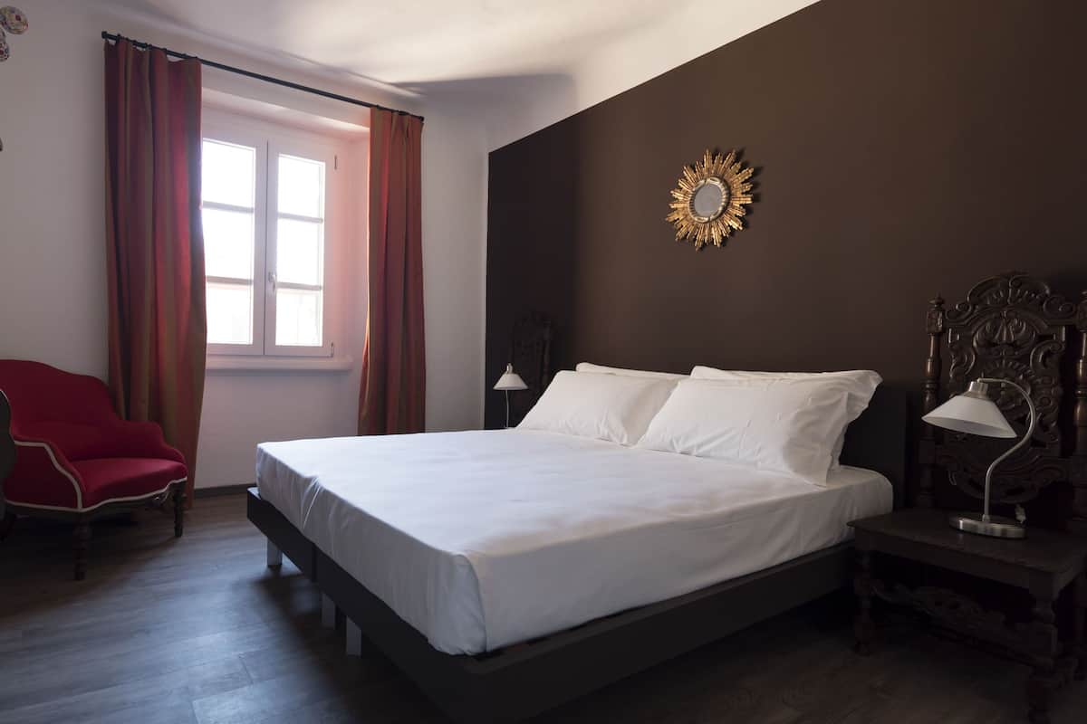 A bedroom with a bed, a red chair, a nightstand with a lamp, and a wall-mounted mirror.